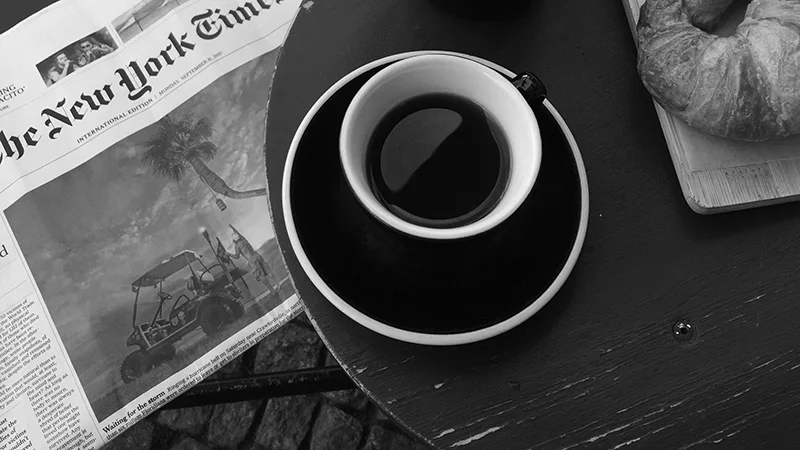 A coffee cup on a table next to a croissant and a newspaper