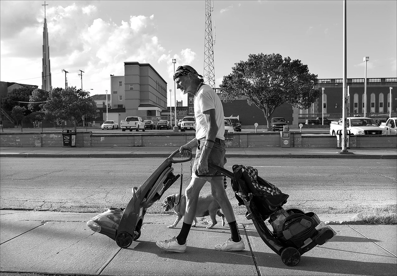 June 2017: Walter and his dog, Blondie, walk down a sidewalk with two vacuum machines Walter recovered from a nearby dumpster. He hopes to sell them. Blondie, like Walter, is homeless. “Blondie’s a good dog. She’s 14 years old. She’s like a 90-something-year-old person so she’s kind of slow getting up. She walks at a pretty good trot. She’s a darn good dog, a little pug beagle. They call a puggie. I call her a puzzle. You don’t never know what she’s gonna act like sometimes she be stubborn as all get out. Other times she be just as loving as you’d want. She’s a good dog, though. One day while Walter was homeless, a passerby noticed Walter and Blondie resting near a convenience store and called 9-1-1 because of his concern for the dog. When the police arrived they told the man that if they confiscated the dog she would risk being euthanized. The man asked Walter if he would allow him to foster Blondie for a while. Walter agreed and after a few weeks Walter found a temporary place to stay and retrieved Blondie. Blondie lives on food scraps Walter saves for her from the soup kitchen. “Food stamps don’t buy Gravy Train. … Blondie, she ain’t worth two cents, but I wouldn’t take $100 for her even though I could use the money real bad.”