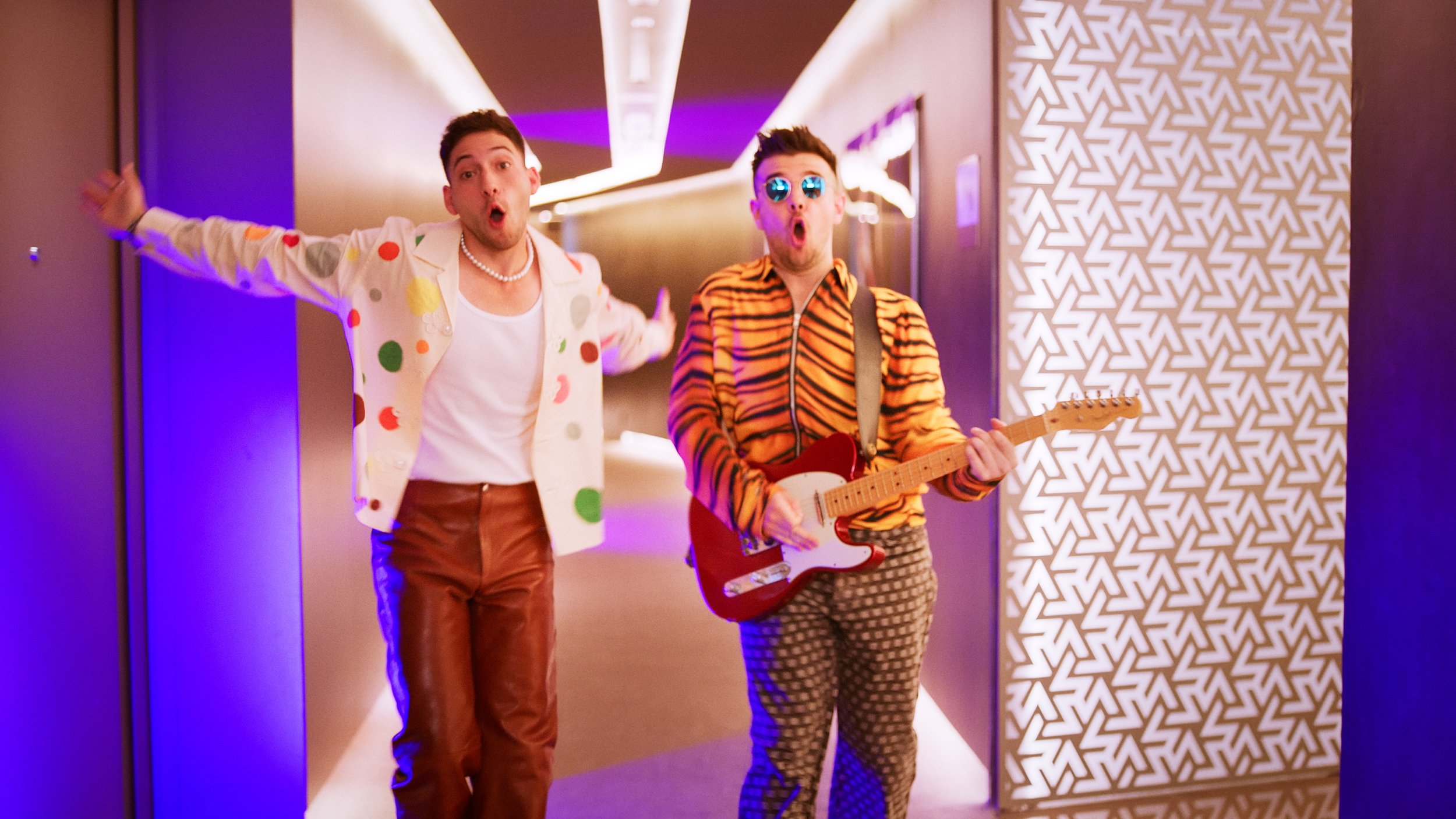 Two individuals in vibrant clothing in a hallway; one holds a guitar. The scene has colorful lighting and patterned walls.