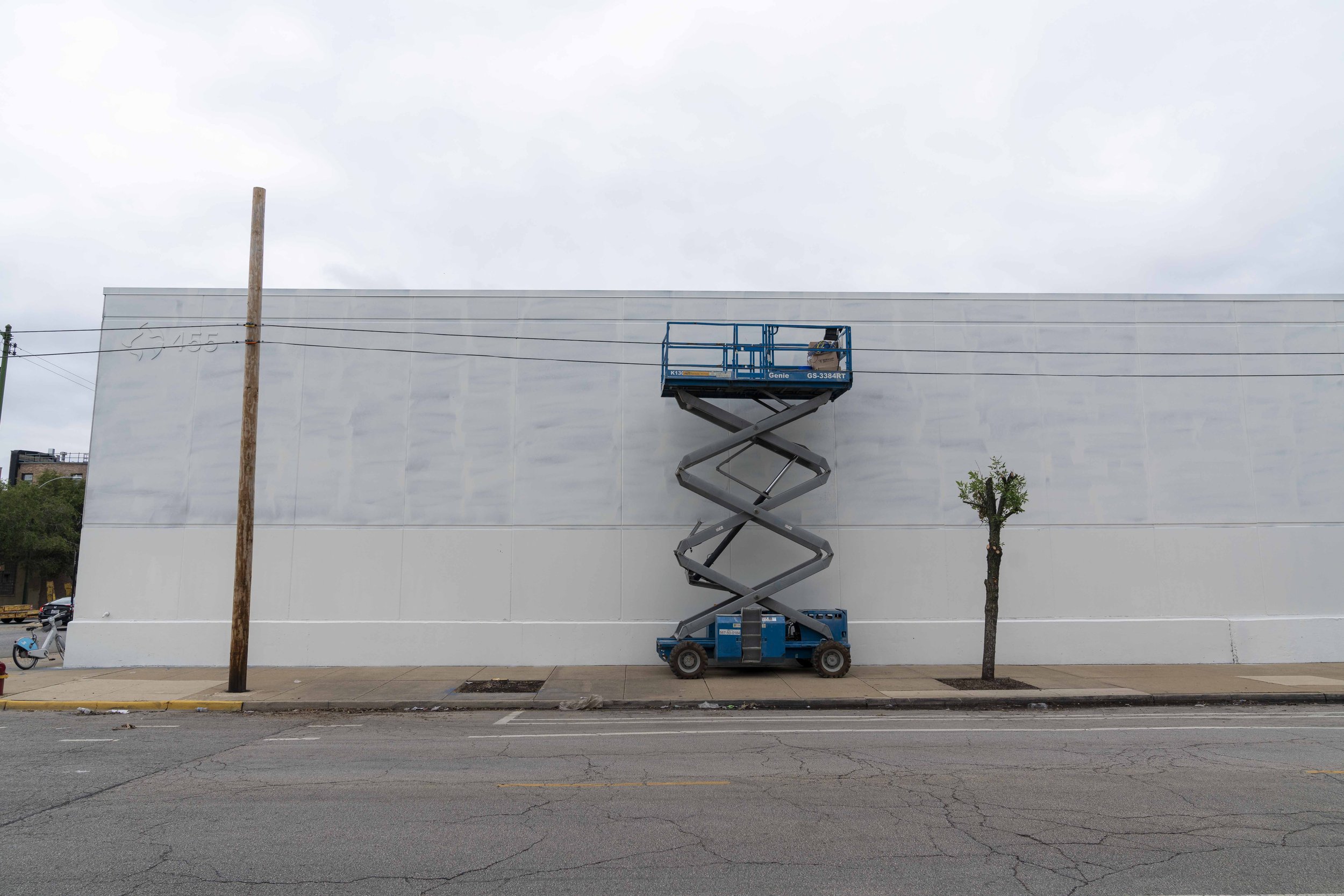 Scissor lift parked beside a white building wall next to a small trimmed tree on a street with a cloudy sky.