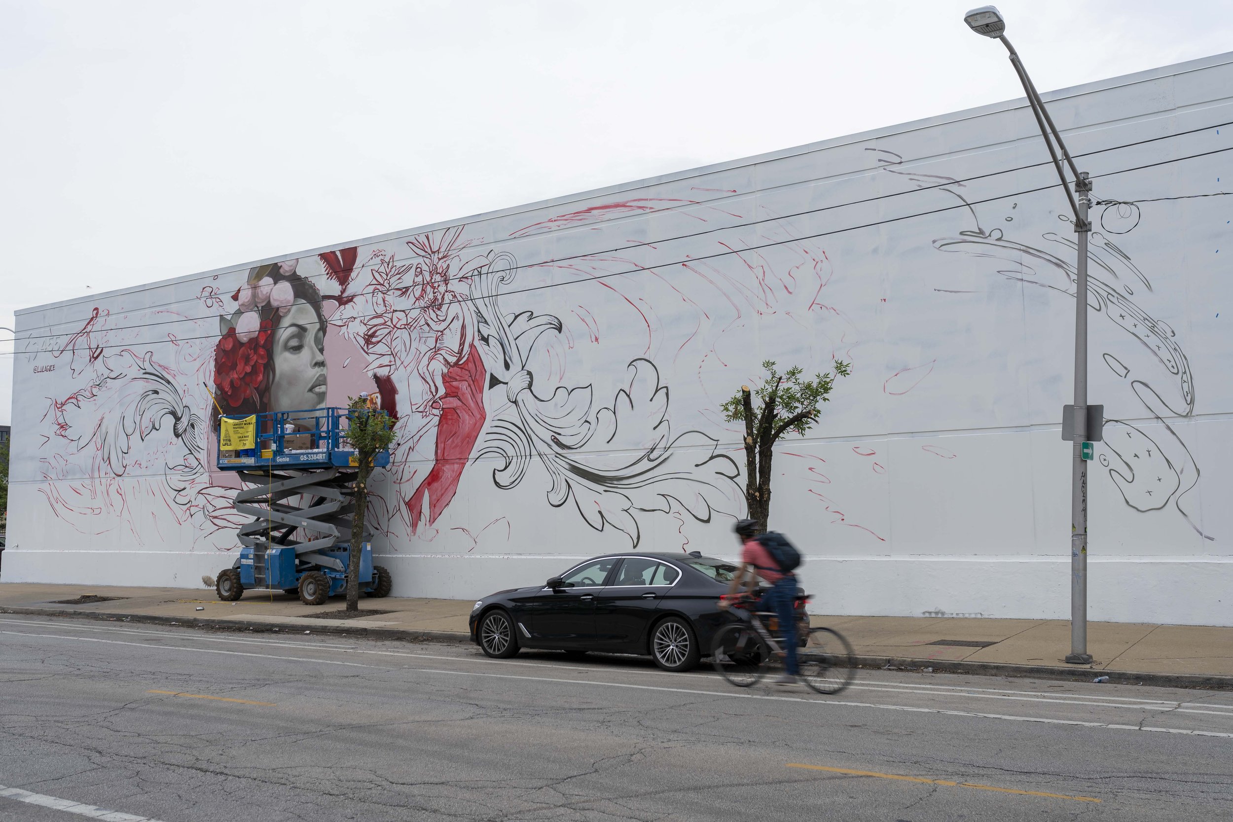 Large mural on a building wall featuring a partially completed portrait of a woman with flowers. An artist's lift is parked nearby. A bicyclist passes by a car on the street in front.
