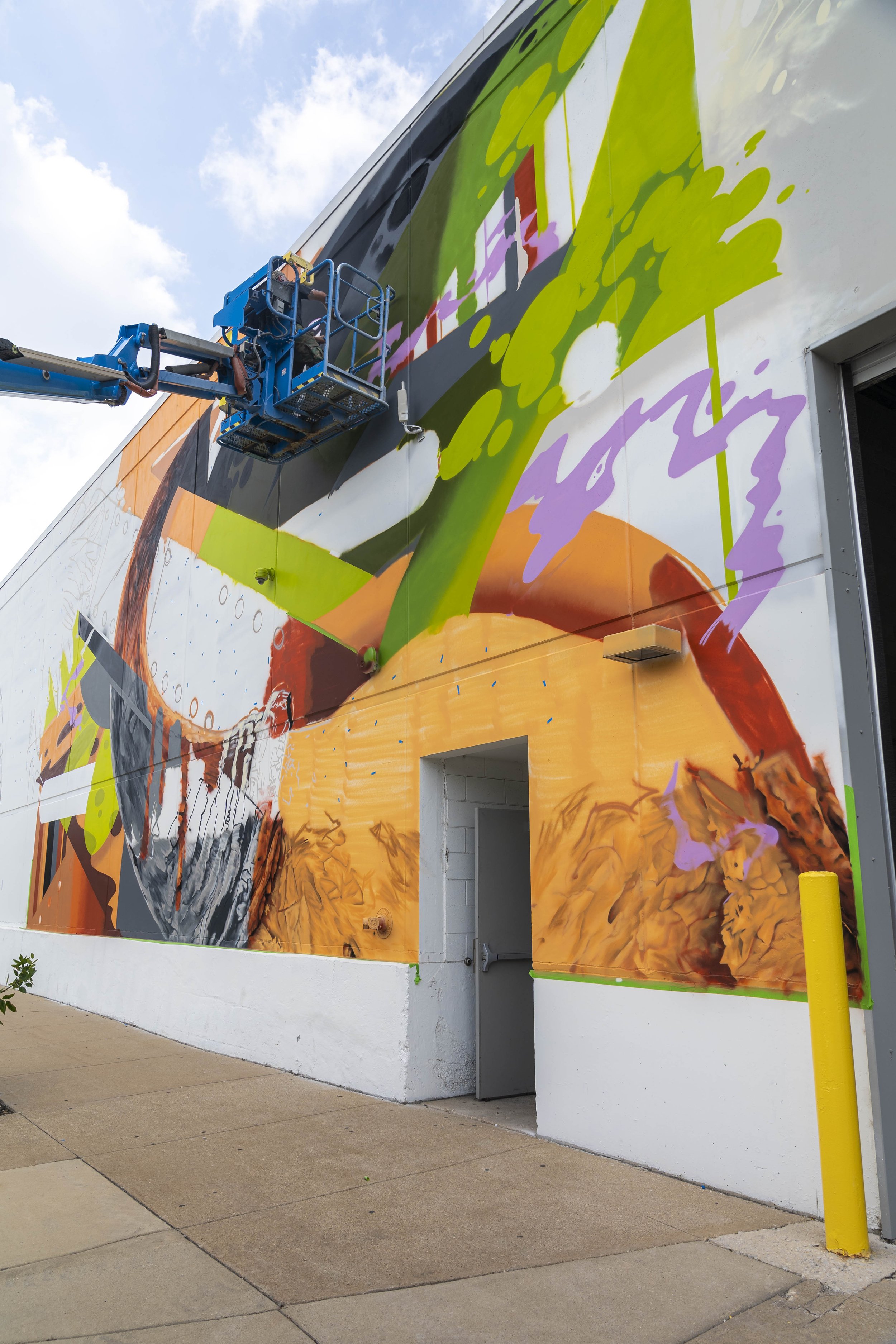 A colorful abstract mural in progress on a large building wall, with a person operating a blue boom lift to paint the upper part.