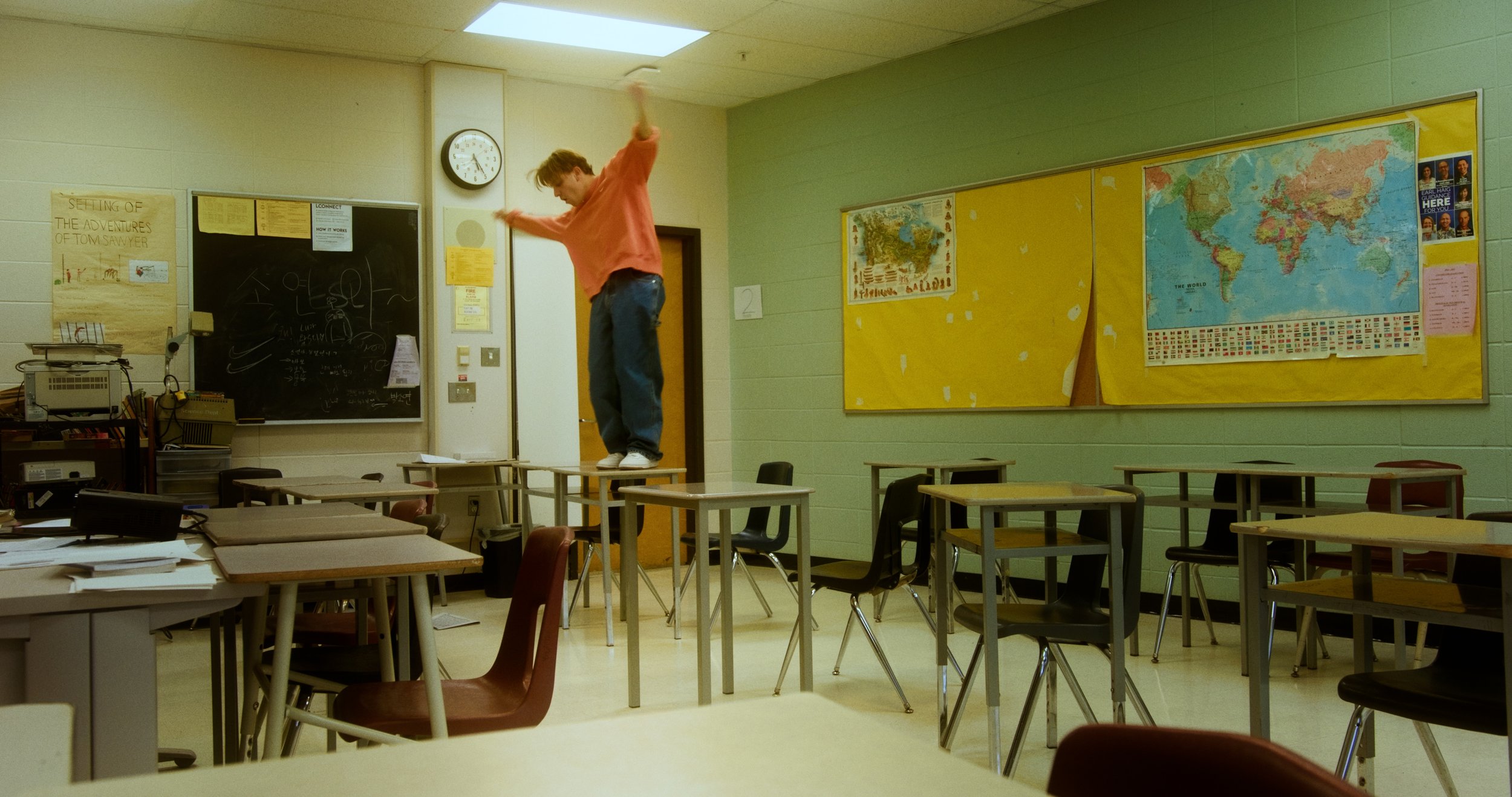 Classroom with empty desks and chairs, wall maps, and a person standing on a desk.