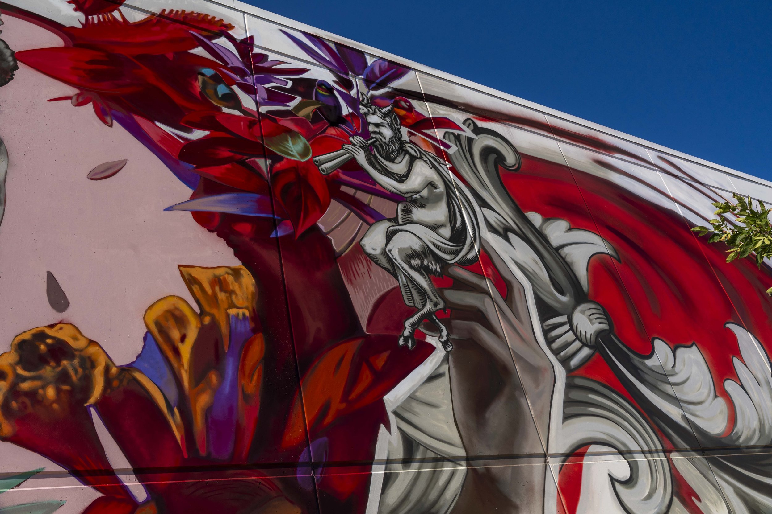 Colorful mural featuring a mythological figure playing a flute, surrounded by vibrant flowers and abstract designs on a wall against a clear blue sky.