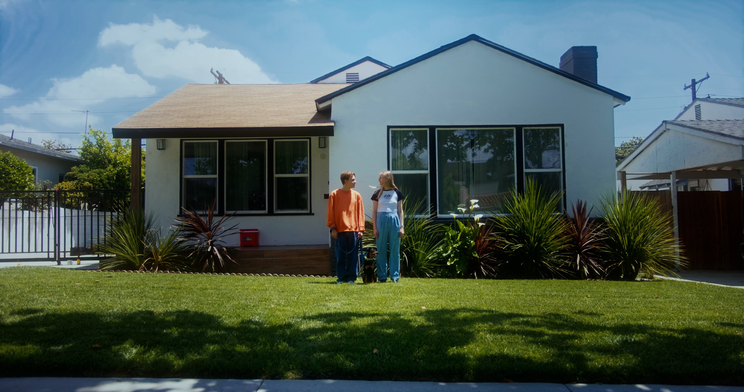 A man and a woman standing on a lawn in front of a white house with large windows and a wooden roof. They are talking and appear relaxed. There are plants and bushes in front of the house, and a clear blue sky above. A dog is sitting beside them.