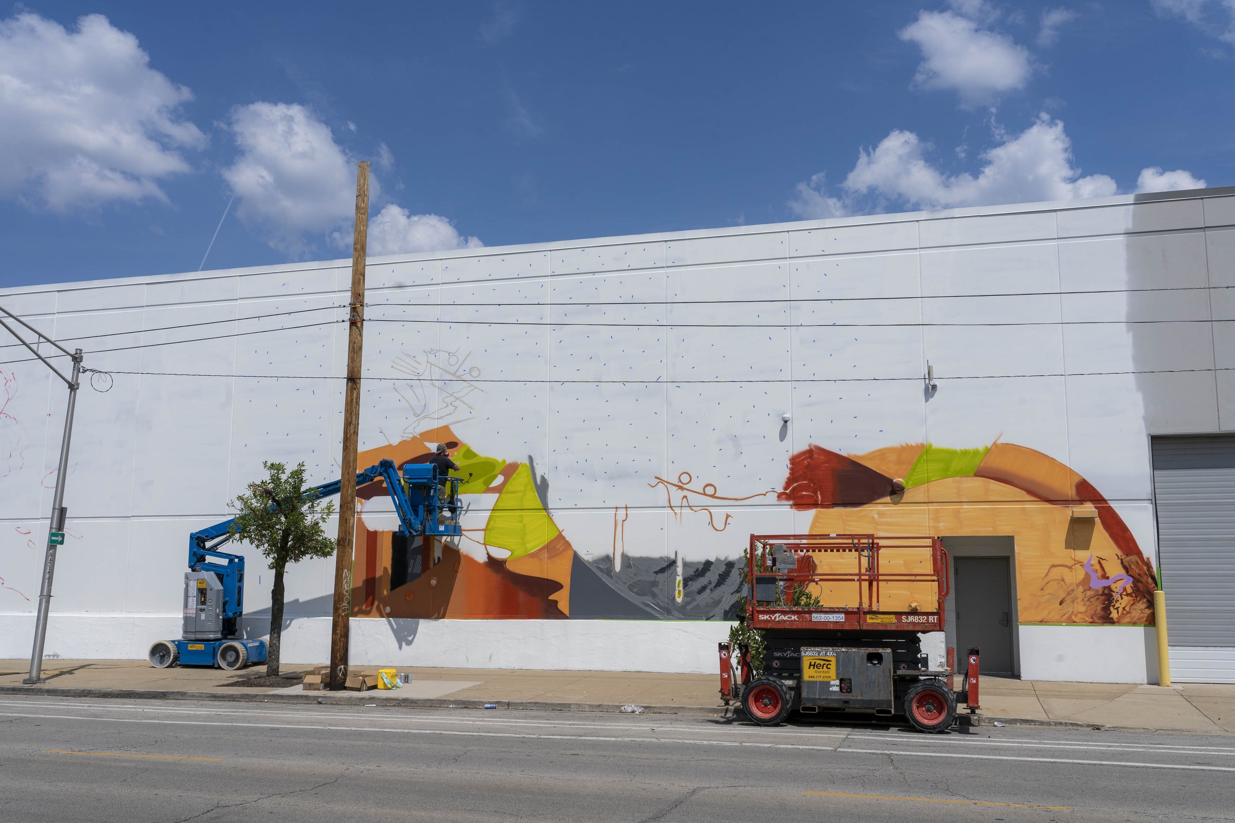 Artists working on a colorful mural on the side of a large building, using hydraulic lifts. The mural features abstract and geometric designs with bright colors. Clear sky and street scene visible.