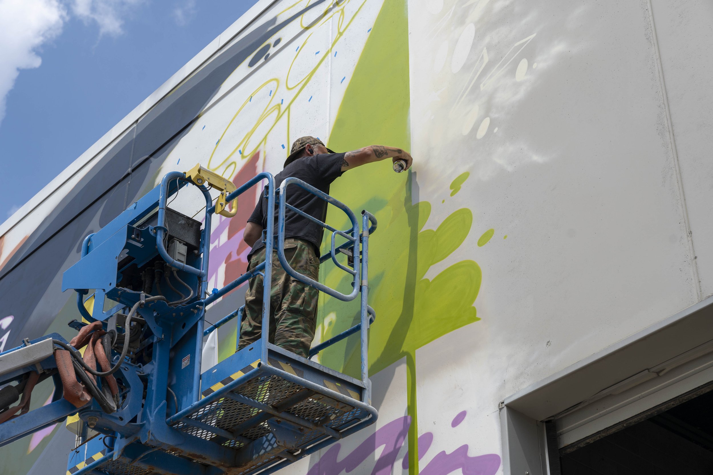 Person painting a large mural on a building using a cherry picker.