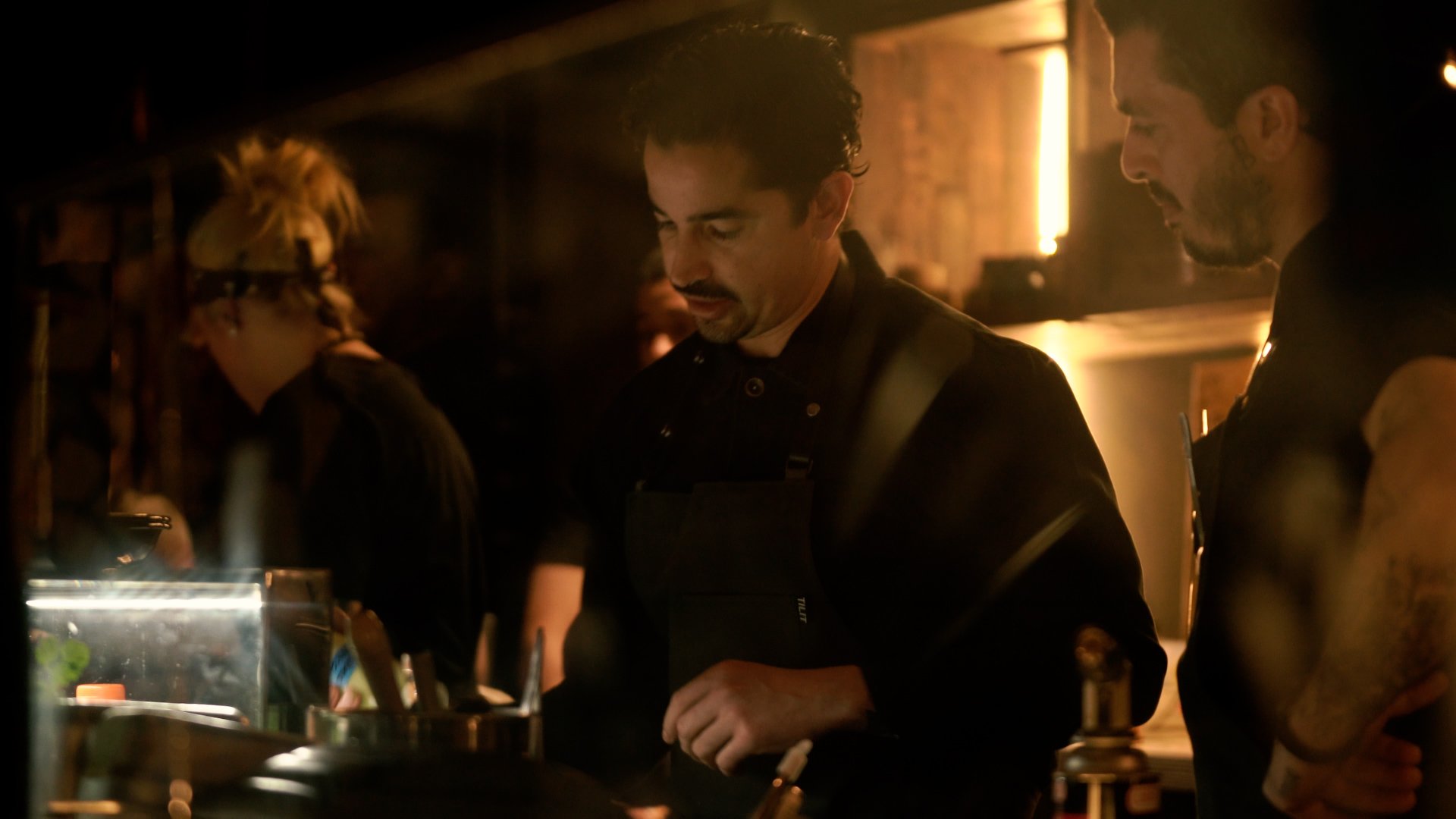 Chefs working in a dimly lit kitchen, wearing black aprons, focused on food preparation.