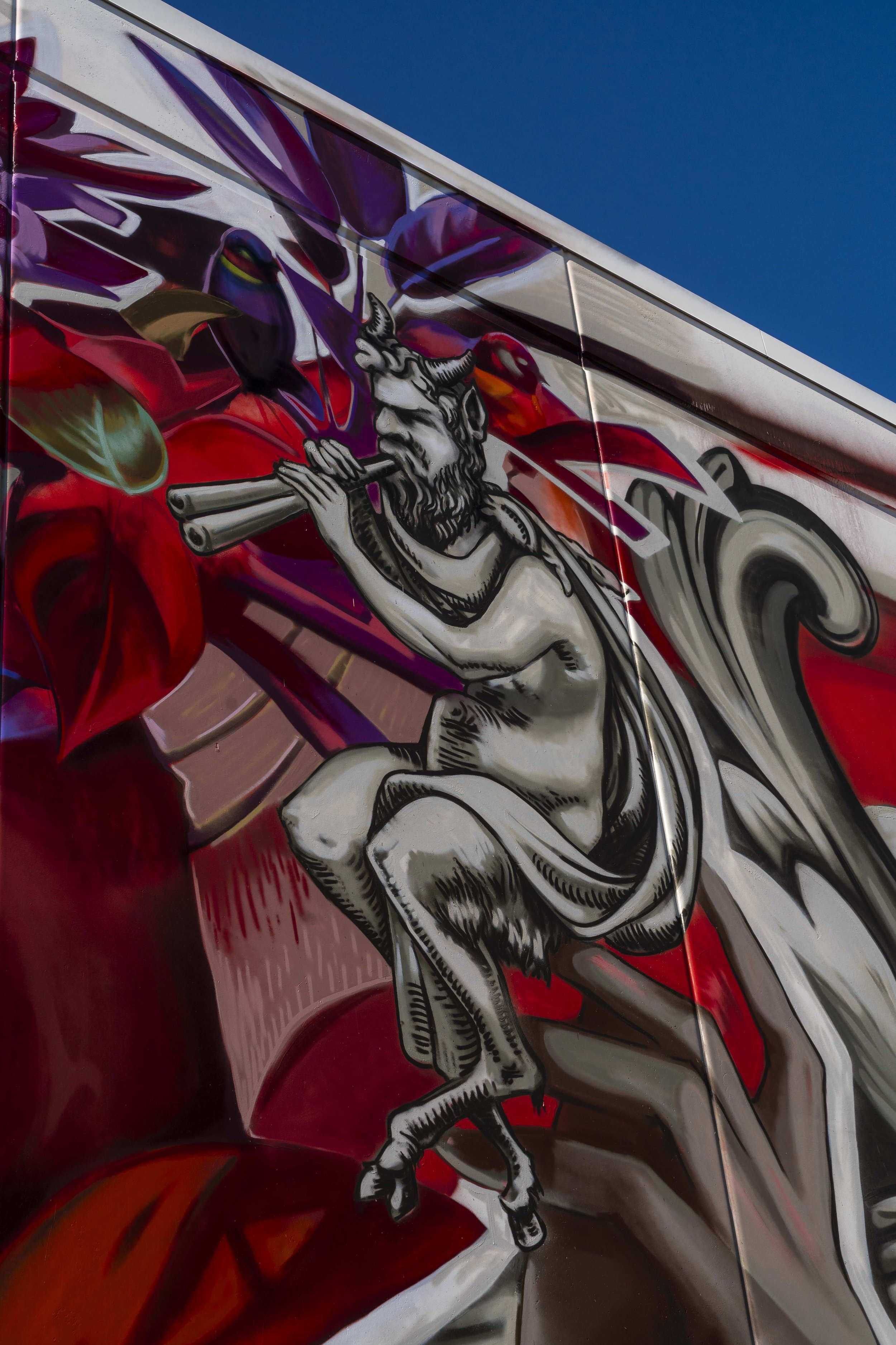 Street art mural featuring a mythical figure playing a flute, surrounded by vibrant red and purple abstract designs against a clear blue sky.