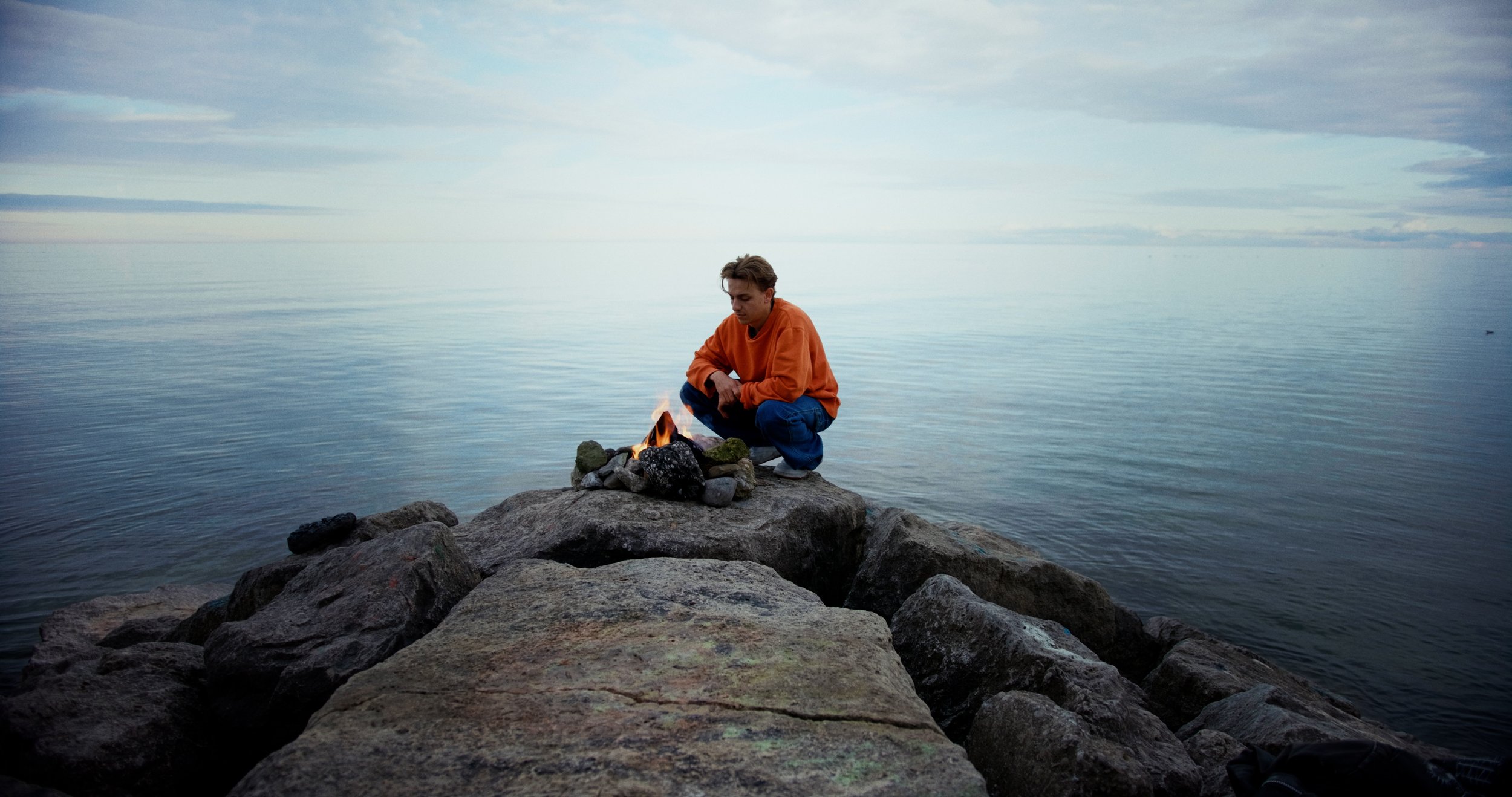 Person in orange sweater sitting by a campfire on a rocky shore, with a calm sea in the background.