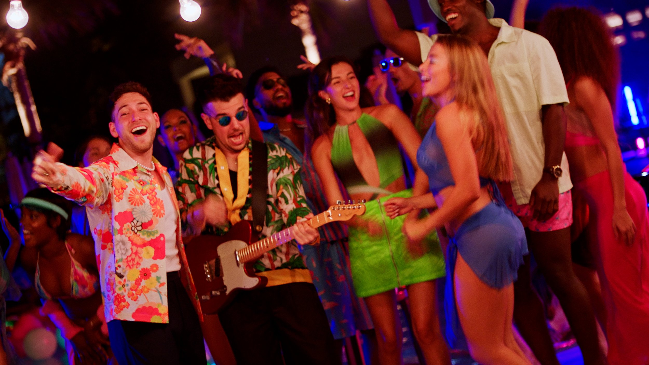 Group of people dancing and having fun at a party with colorful outfits, one person playing a guitar, under dim lights.