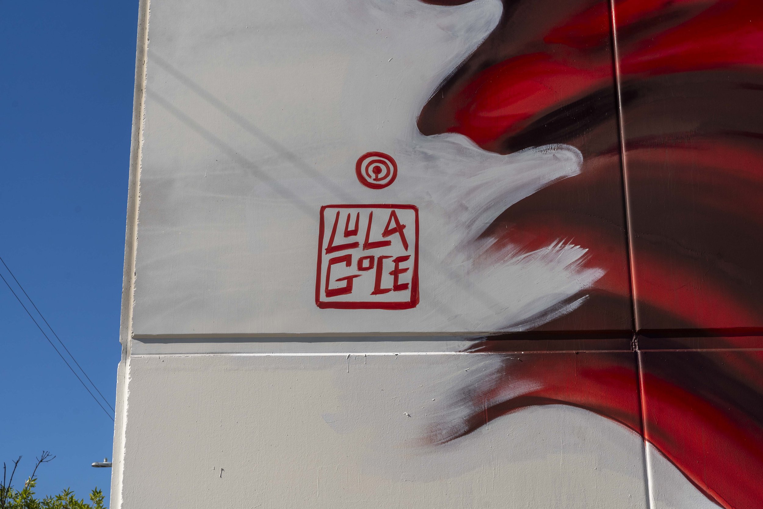 Street art mural featuring abstract red and white swirls with a small, red square signature reading 'Lula Goce' on a white wall under a clear blue sky.