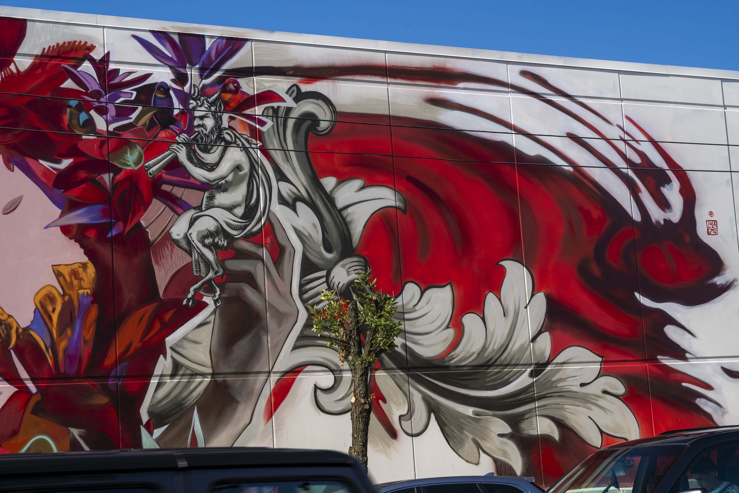 Street mural depicting a mythological figure playing a flute, surrounded by vibrant red and purple abstract designs on a large wall.