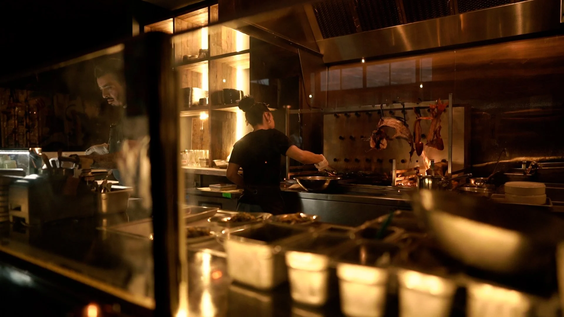 Chefs working in a professional kitchen with subdued lighting, preparing food on a stovetop and using various cookware.
