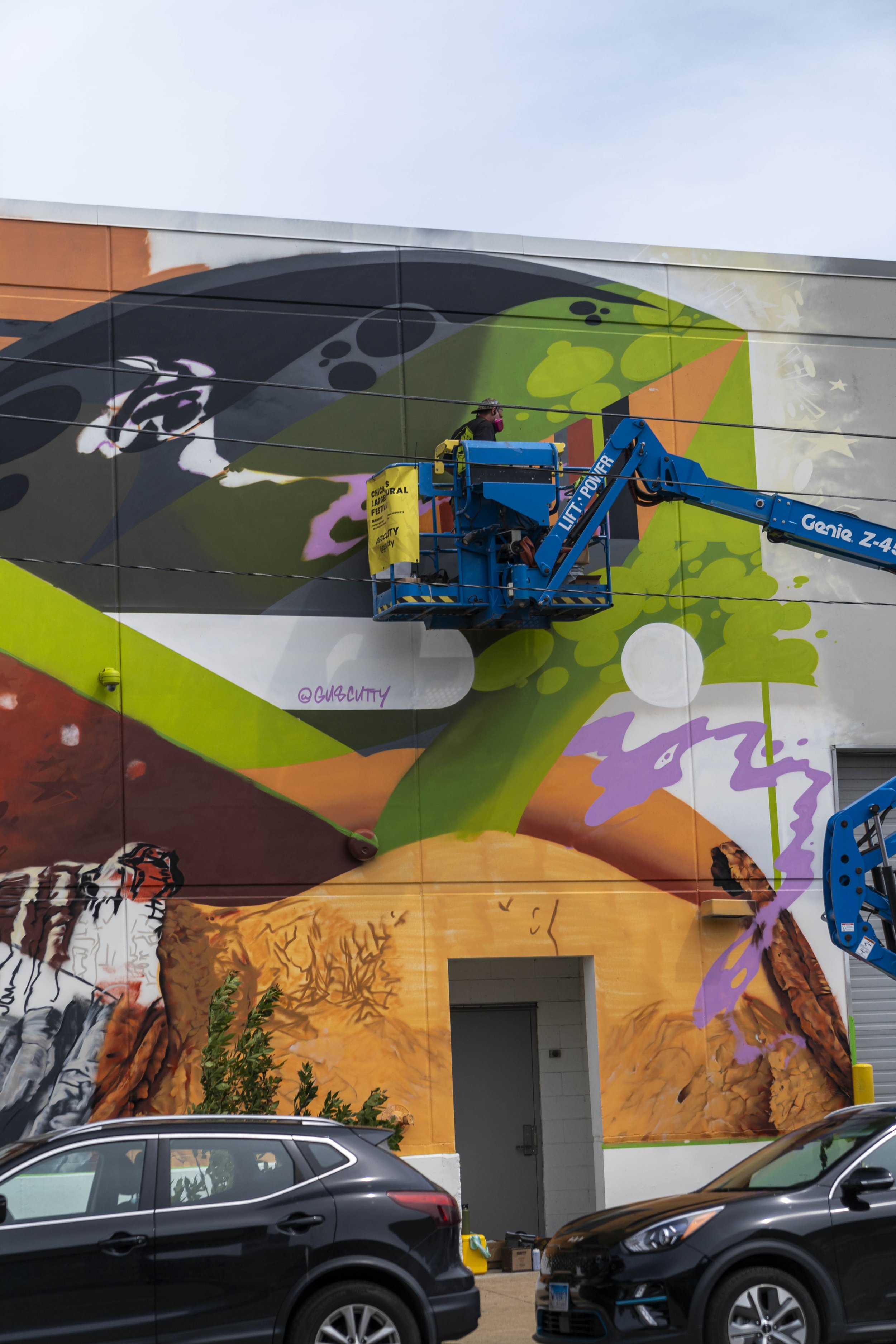 Artist working on colorful mural on building wall with lift, featuring abstract and natural elements.
