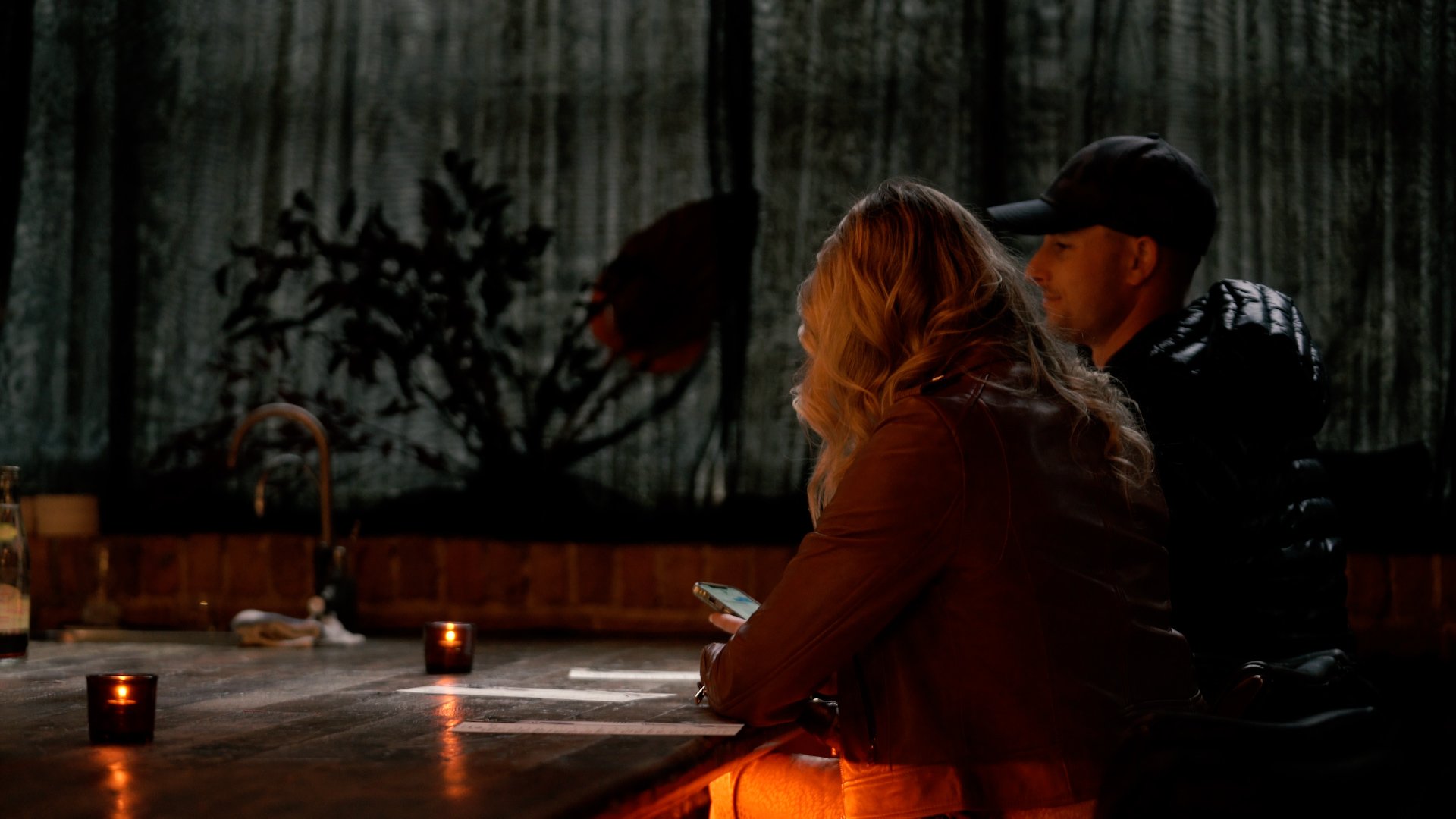 A dimly lit bar scene with a woman and man sitting at the counter. The woman is using a smartphone, and there are candles on the counter. The background is dark with silhouetted plants or decorations.