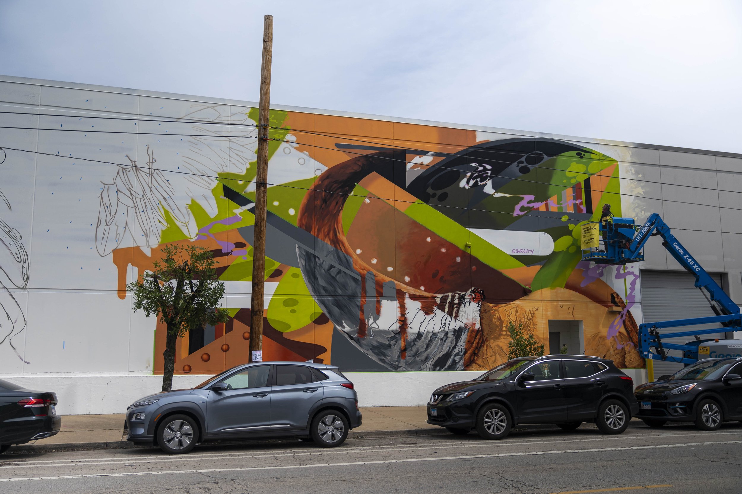 Street art mural on a building wall with abstract colorful shapes, a person on a lift painting, cars parked in front, and a tree nearby.