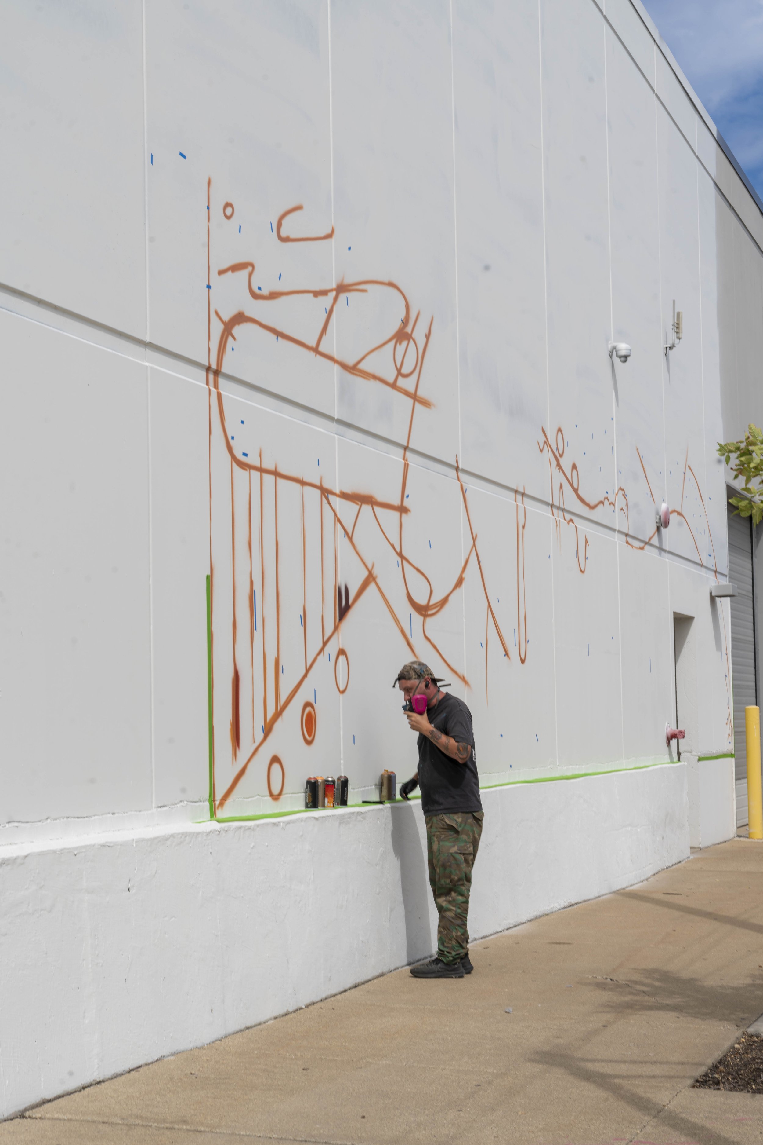 Artist preparing to paint a mural on a large white wall, with initial sketch lines and spray paint cans visible.