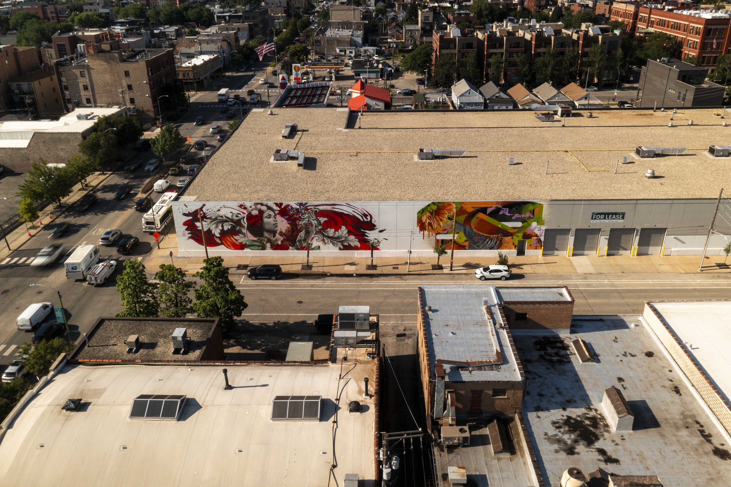 Aerial view of an urban area with a large building featuring colorful murals on its side, depicting artistic faces and designs. Streets with vehicles and trees surround the building. The area includes various residential and commercial structures.