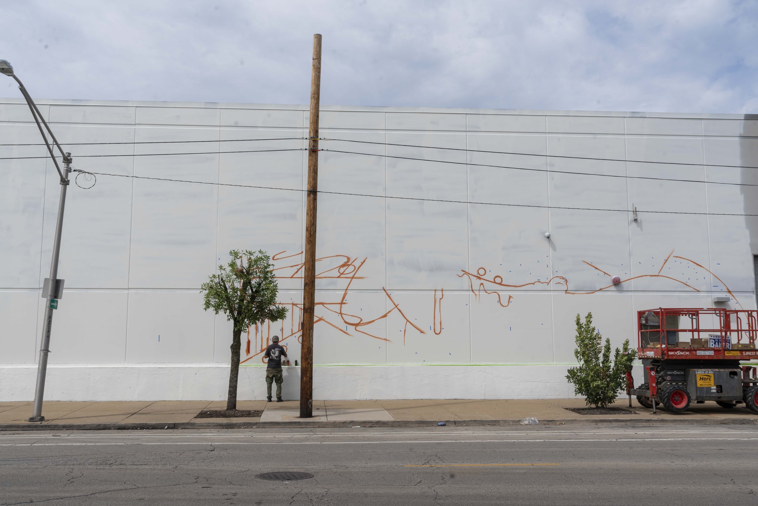 Person painting outlines for a mural on a large white wall, with a tree and utility pole in front. A lift platform is parked nearby on the sidewalk.