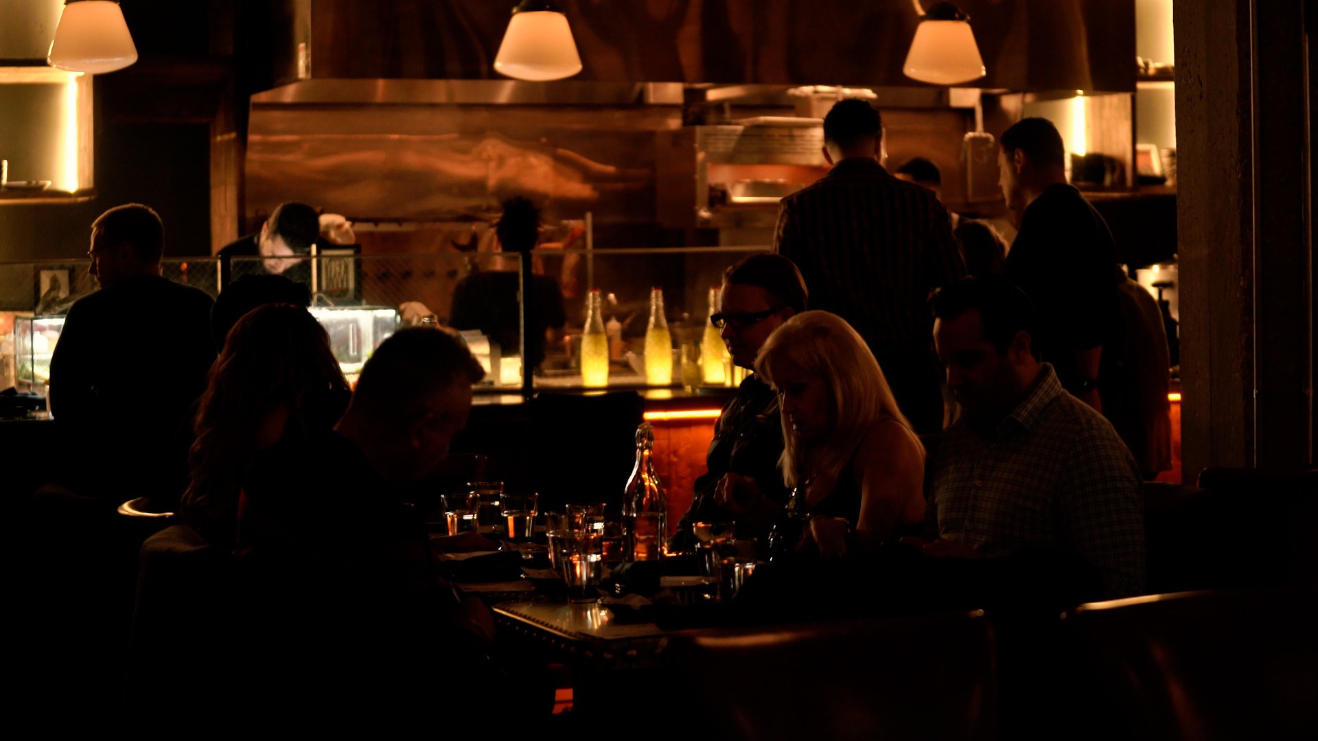 Dimly lit restaurant interior with patrons seated at tables and chefs working behind the counter.