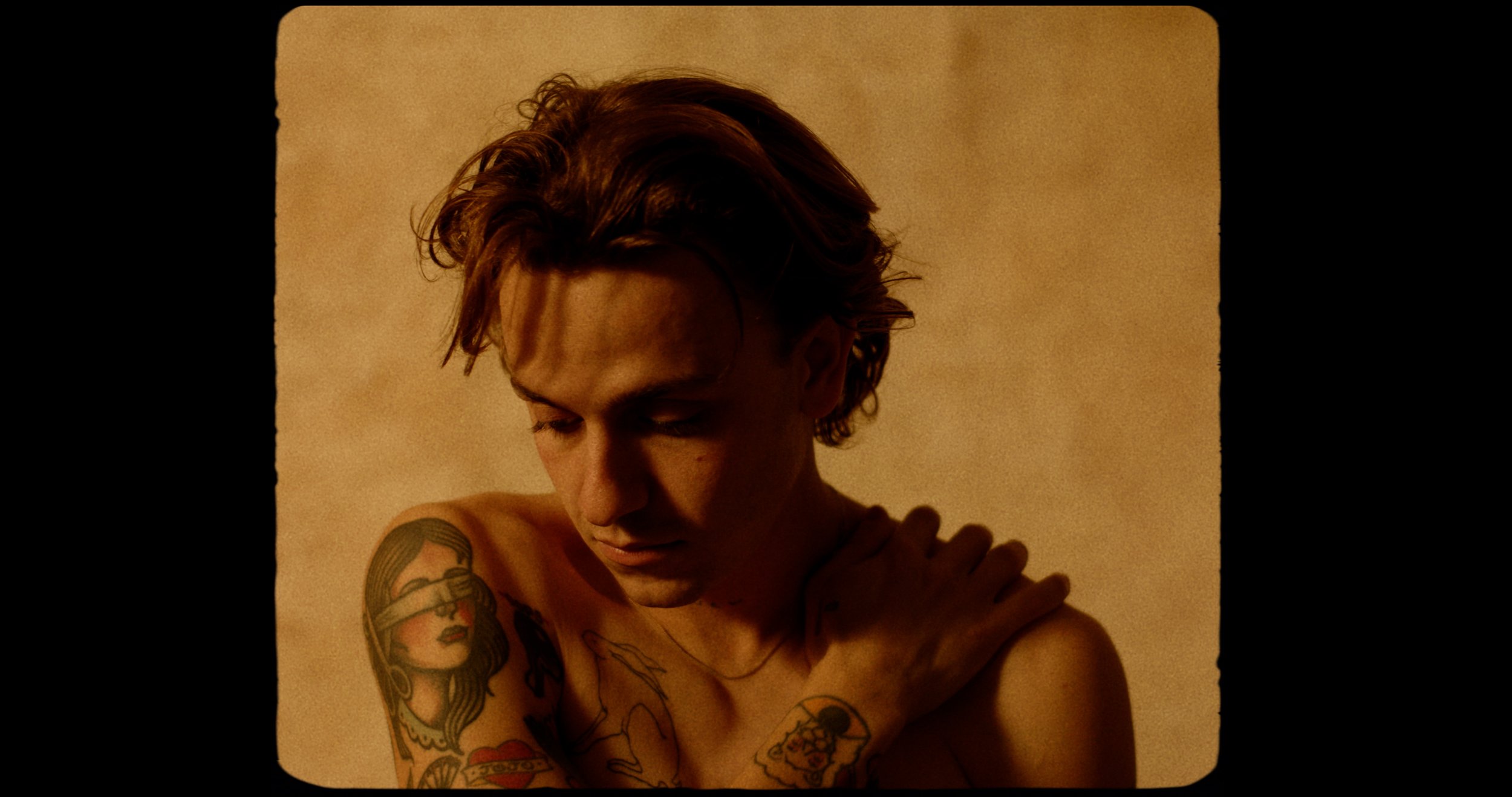 Man with tattoos on arm, looking down, hand on shoulder against a beige background.