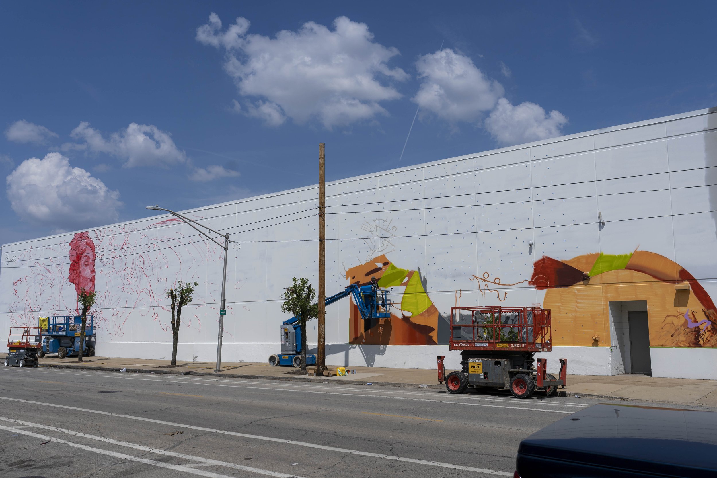 Artists working on a large mural on the side of a building, featuring a partially painted colorful design and a red portrait sketch, using lifts for access.