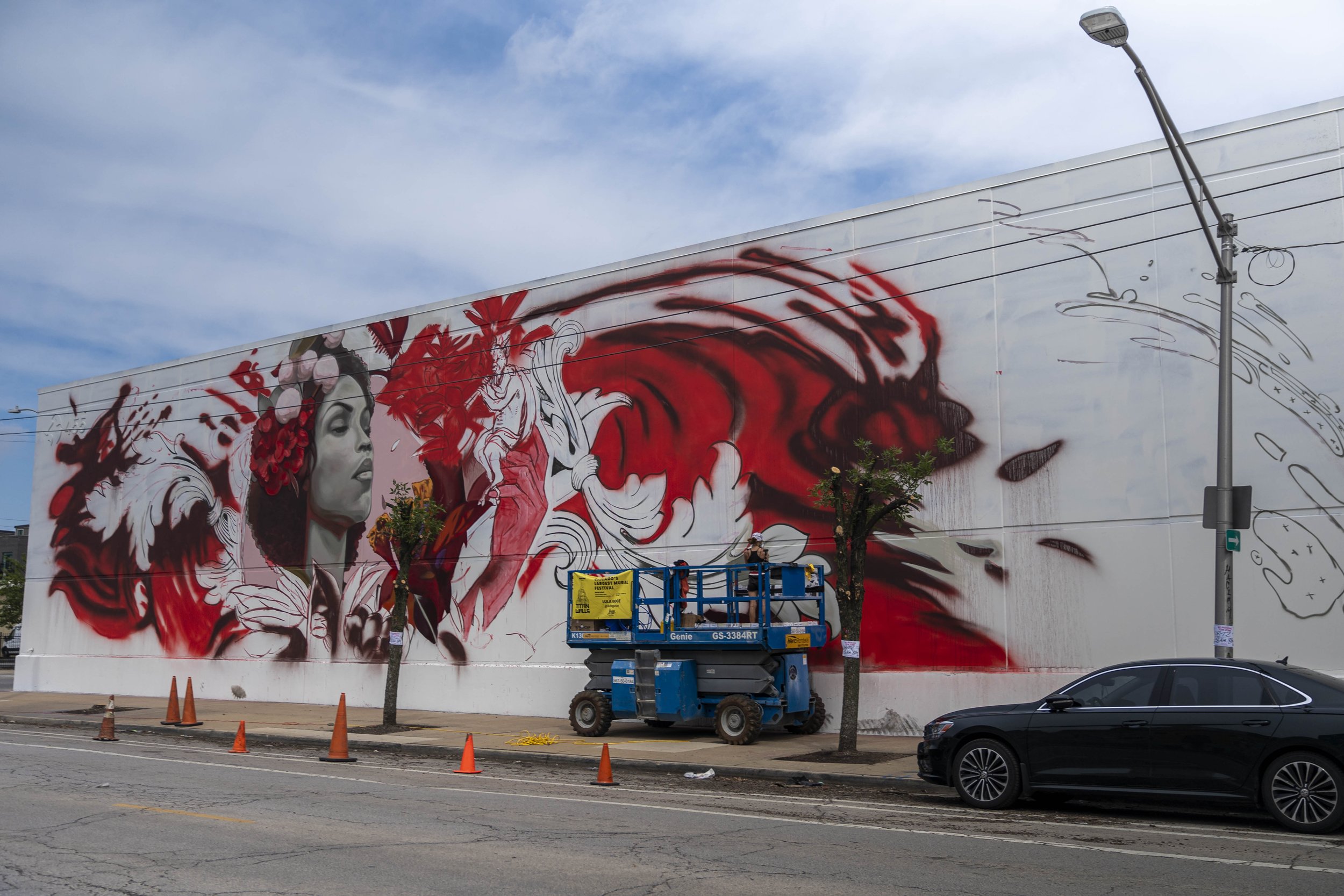 Street art mural depicting a woman's face with red and white abstract patterns on a building; a worker is on a lift in front of the mural, with traffic cones and a black car nearby.