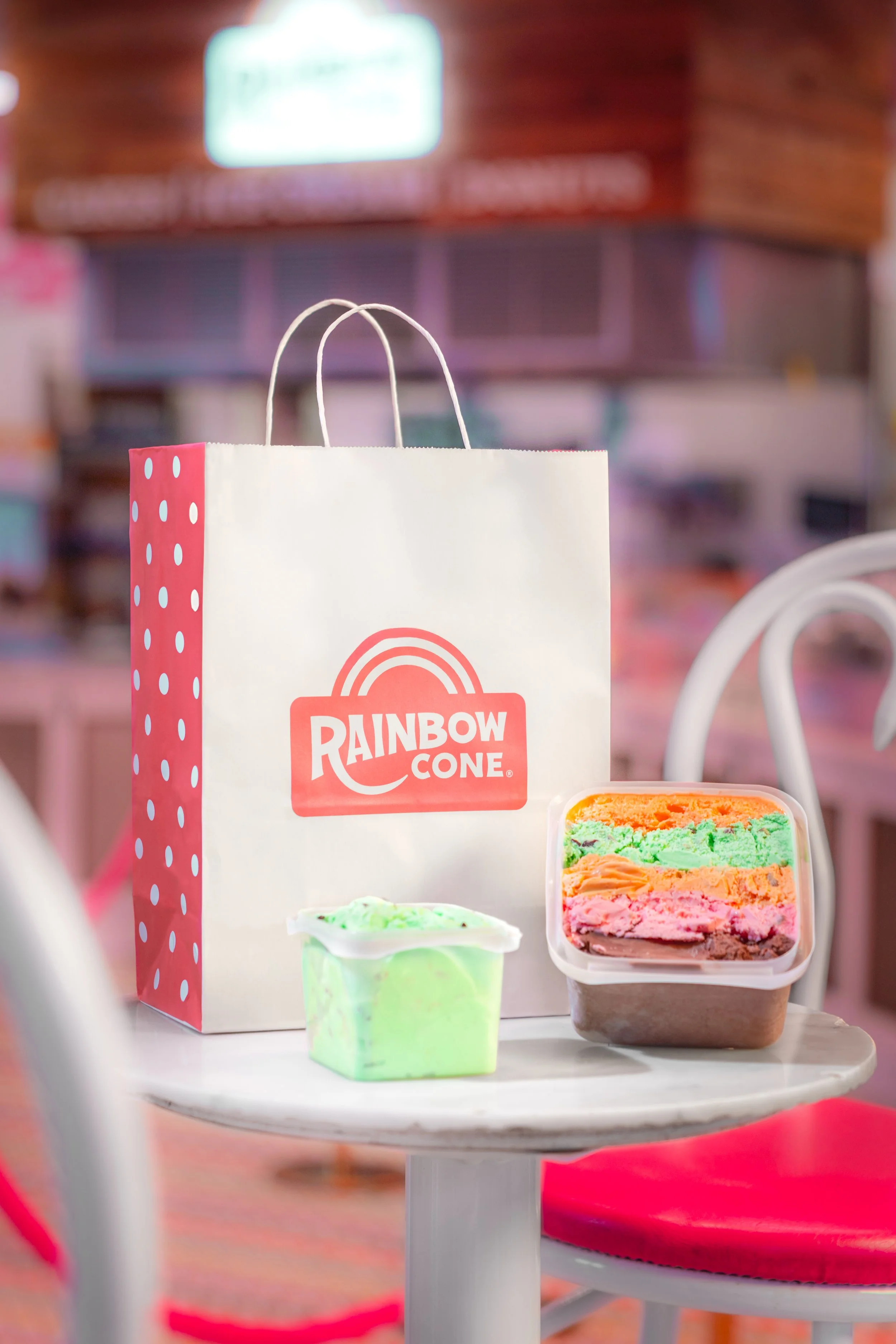 A Rainbow Cone shopping bag and two containers with colorful ice cream on a white table inside an ice cream shop.