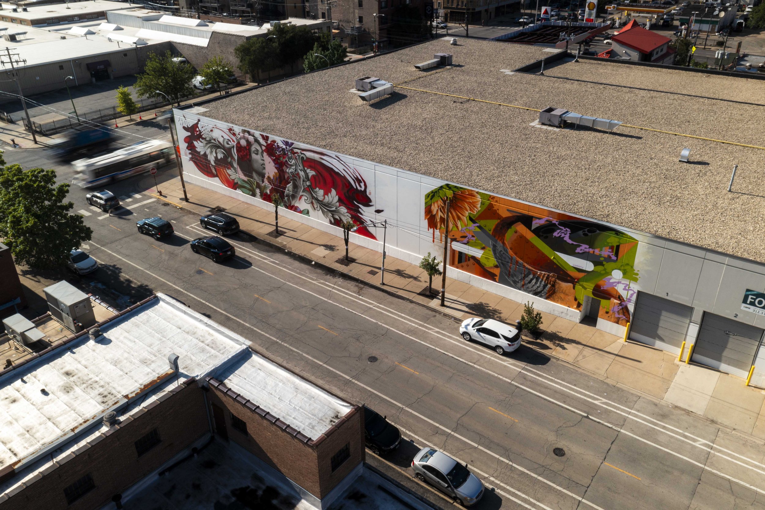 Aerial view of a building with colorful murals on the side, featuring abstract and detailed designs, including a woman's face and floral elements, alongside a street with parked cars and moving vehicles.
