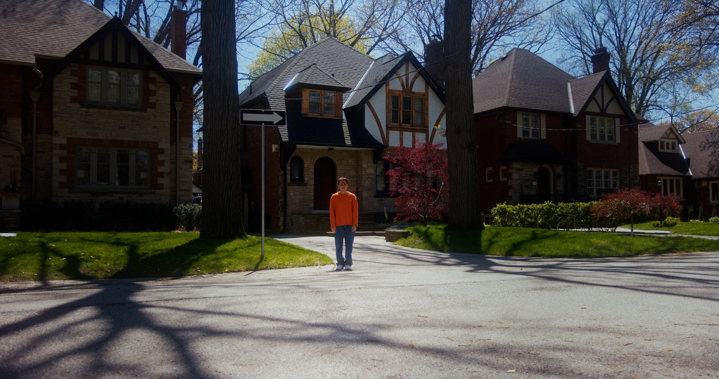 Person in an orange shirt standing on a residential street with Tudor-style houses and trees