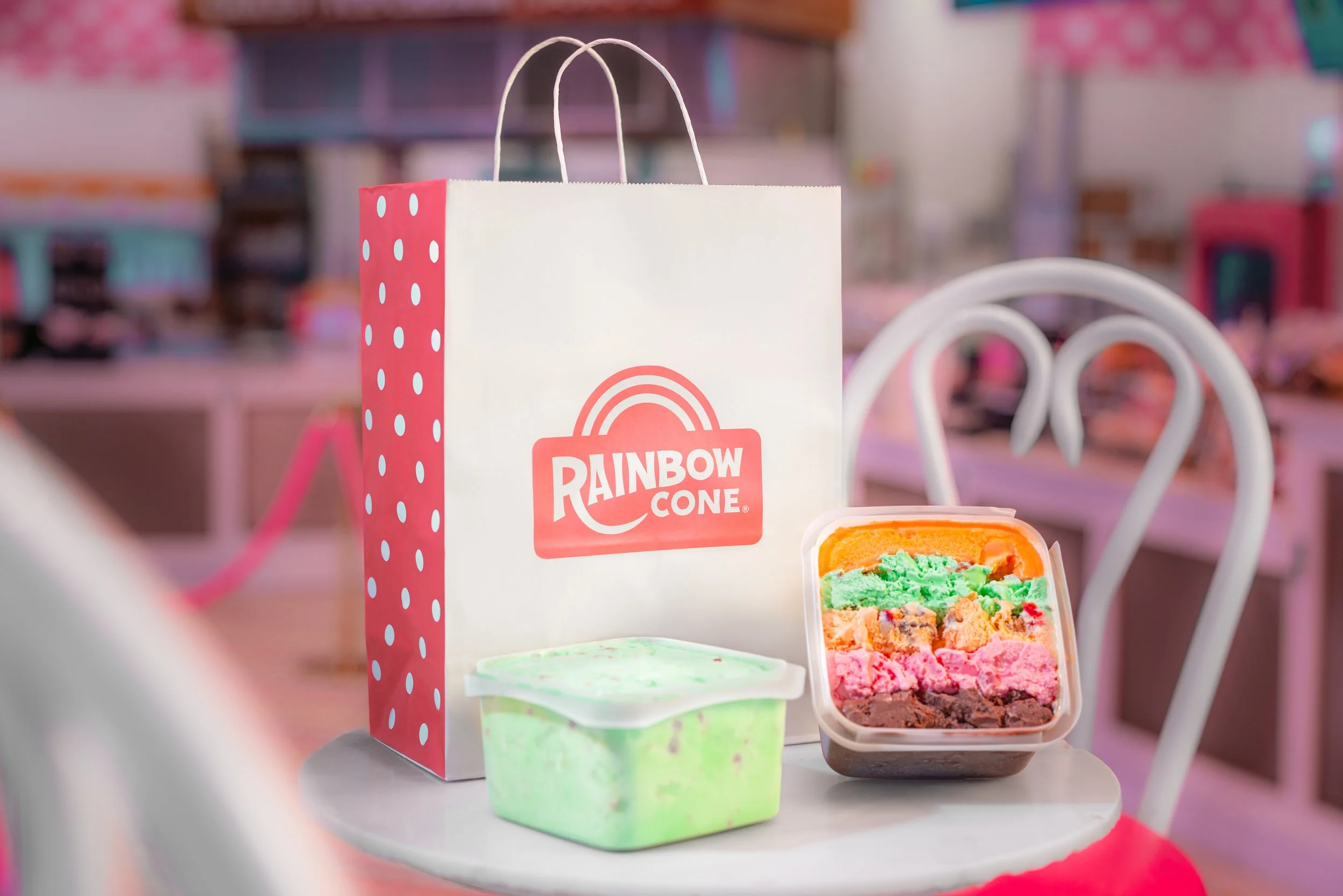 Rainbow Cone shopping bag, a container of rainbow-colored ice cream, and a green ice cream container on a small white table in a pink-themed ice cream shop