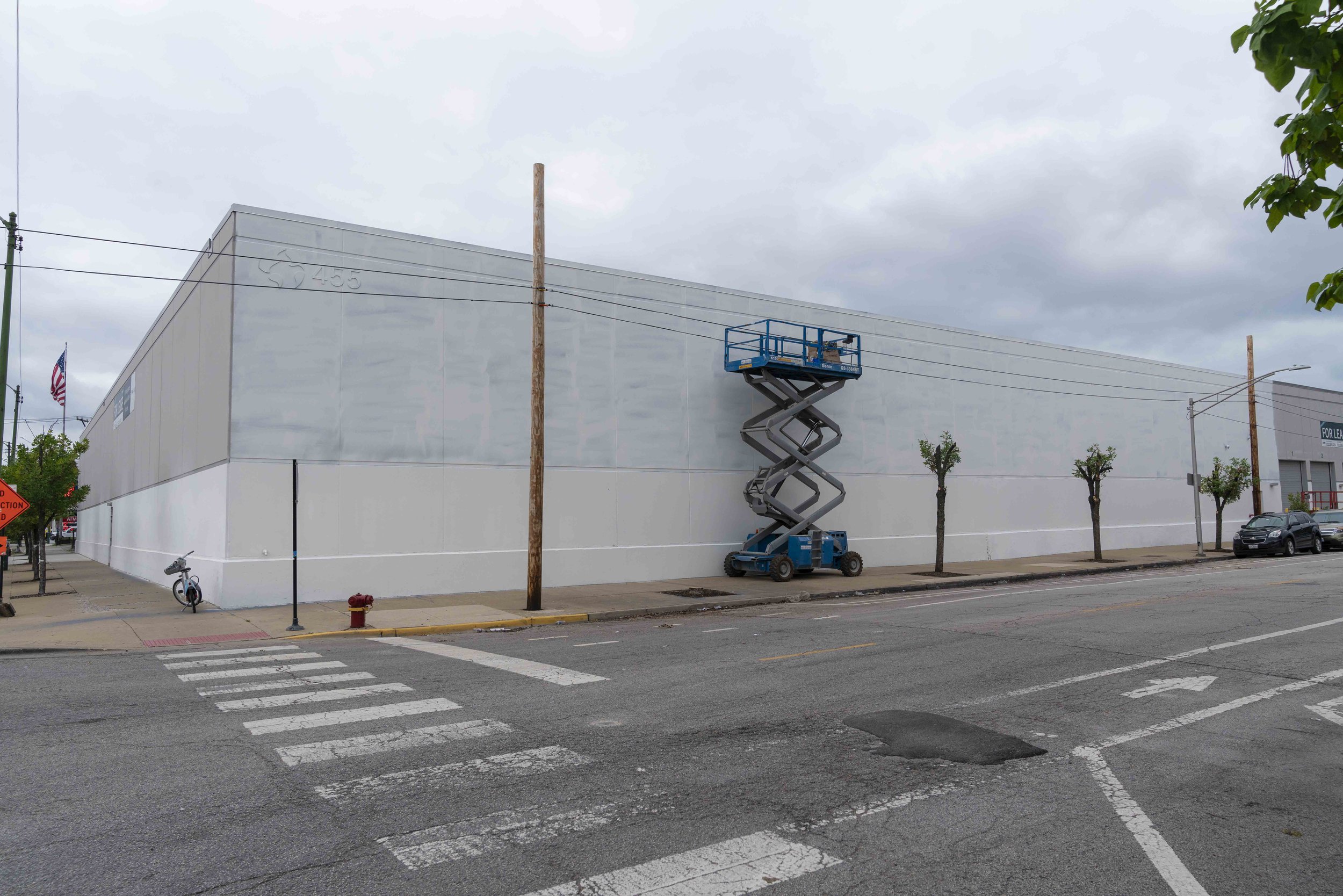 A large industrial building with a gray and white exterior, featuring an elevated scissor lift on the sidewalk. The street in front has crosswalk markings, an orange construction sign, a bicycle, and an American flag in the background.