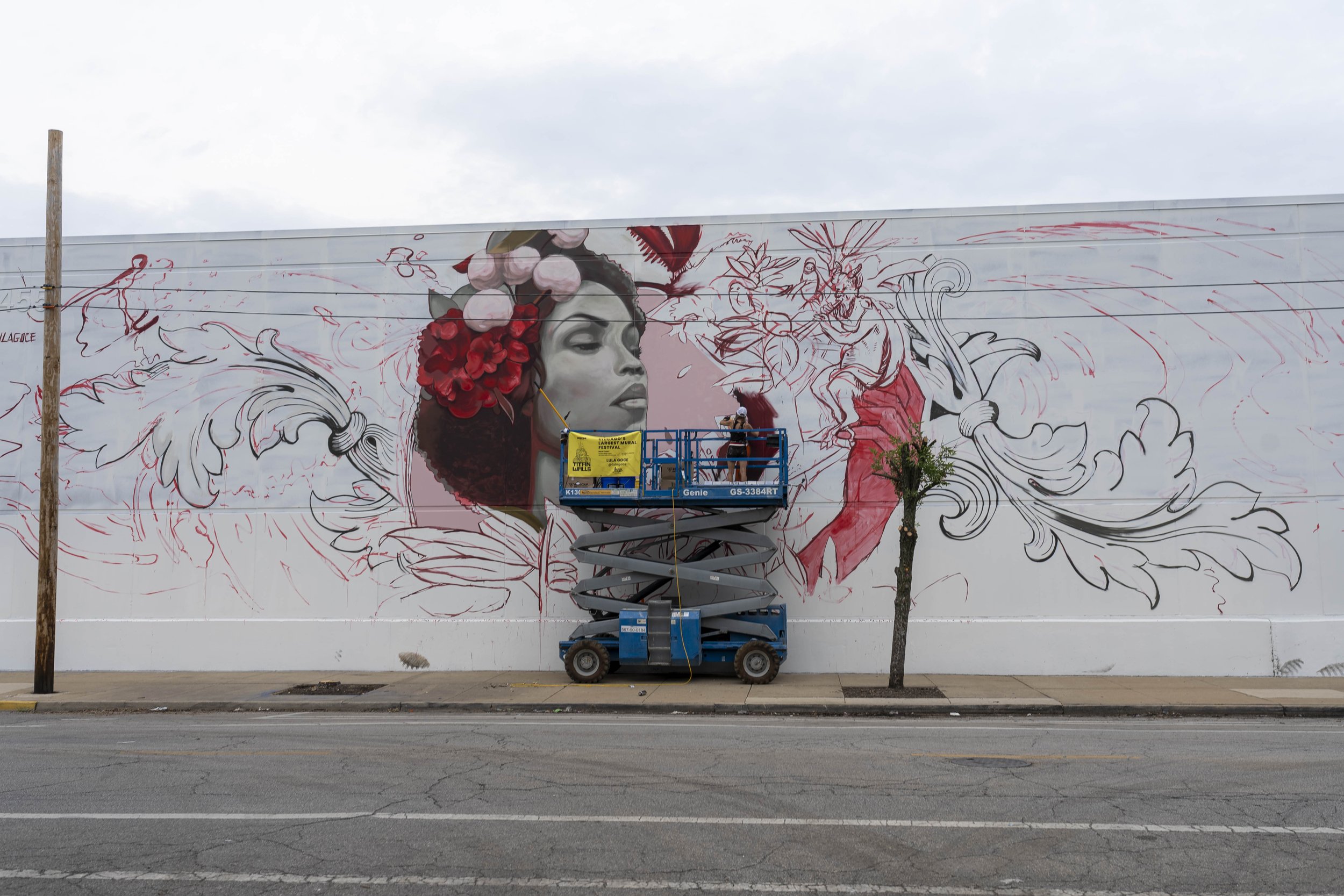 Artist painting a large mural on a wall using a hydraulic lift, featuring a woman's face with floral and abstract designs.