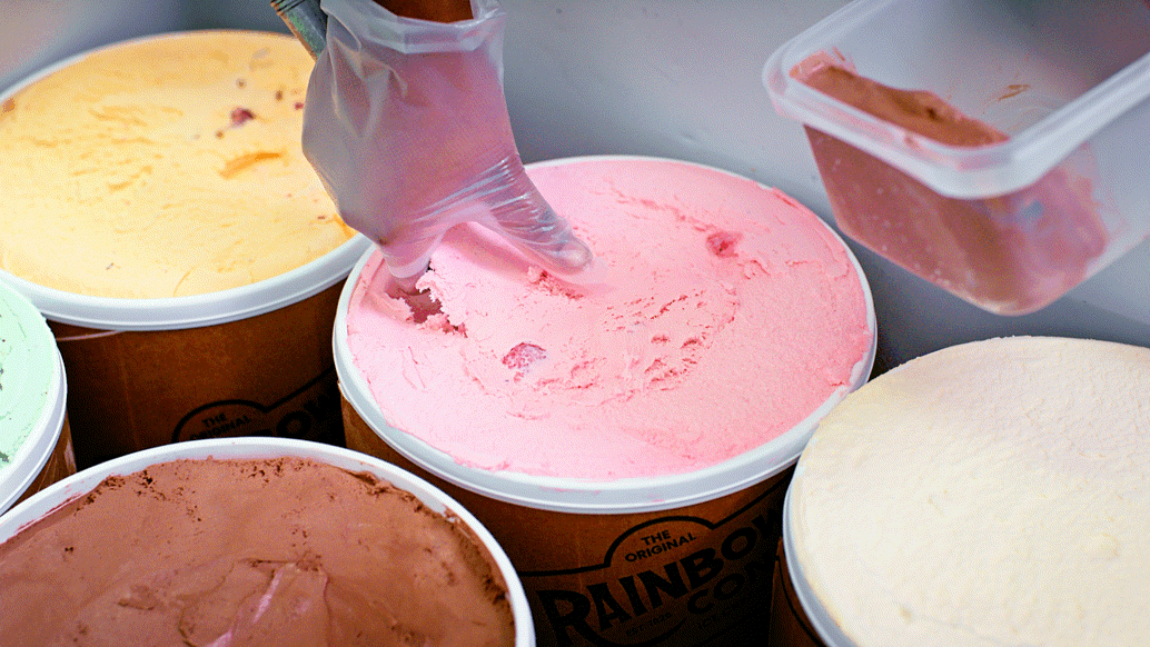 Scoops of colorful ice cream flavors in tubs, with a hand in a glove scooping pink ice cream.