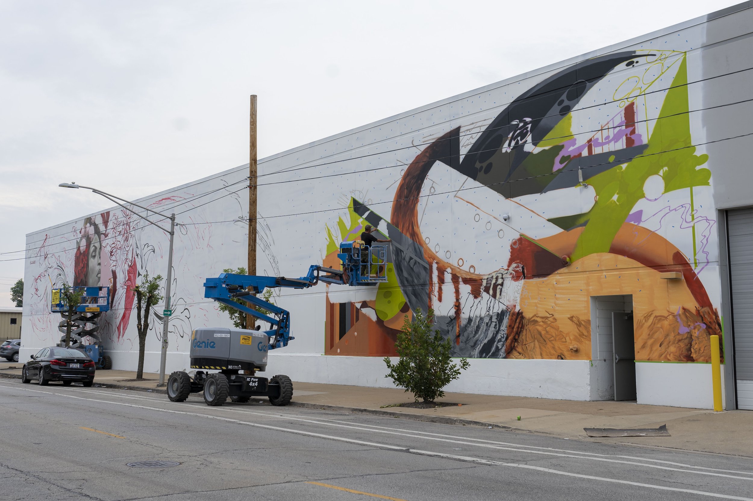 Artists painting a large mural on the side of a building using scissor lifts. The mural features colorful abstract shapes and partial portraits.