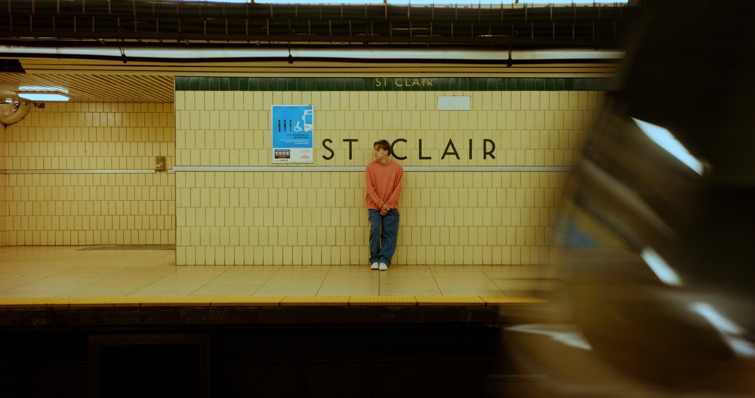 Person in red sweater standing on subway platform at St. Clair station, train passing in foreground.