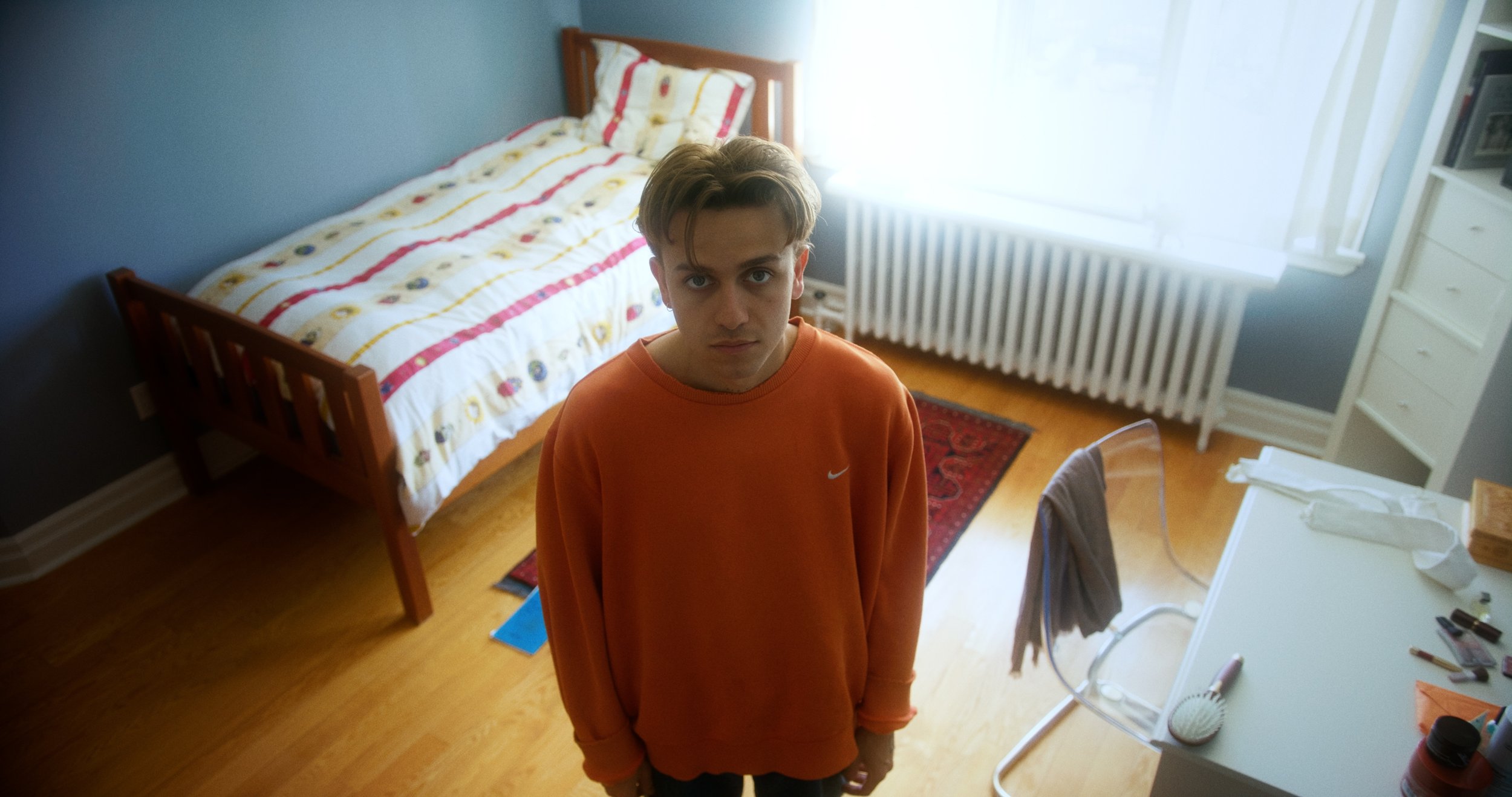 Young person in an orange sweater standing in a tidy bedroom with a bed, desk, and chair.
