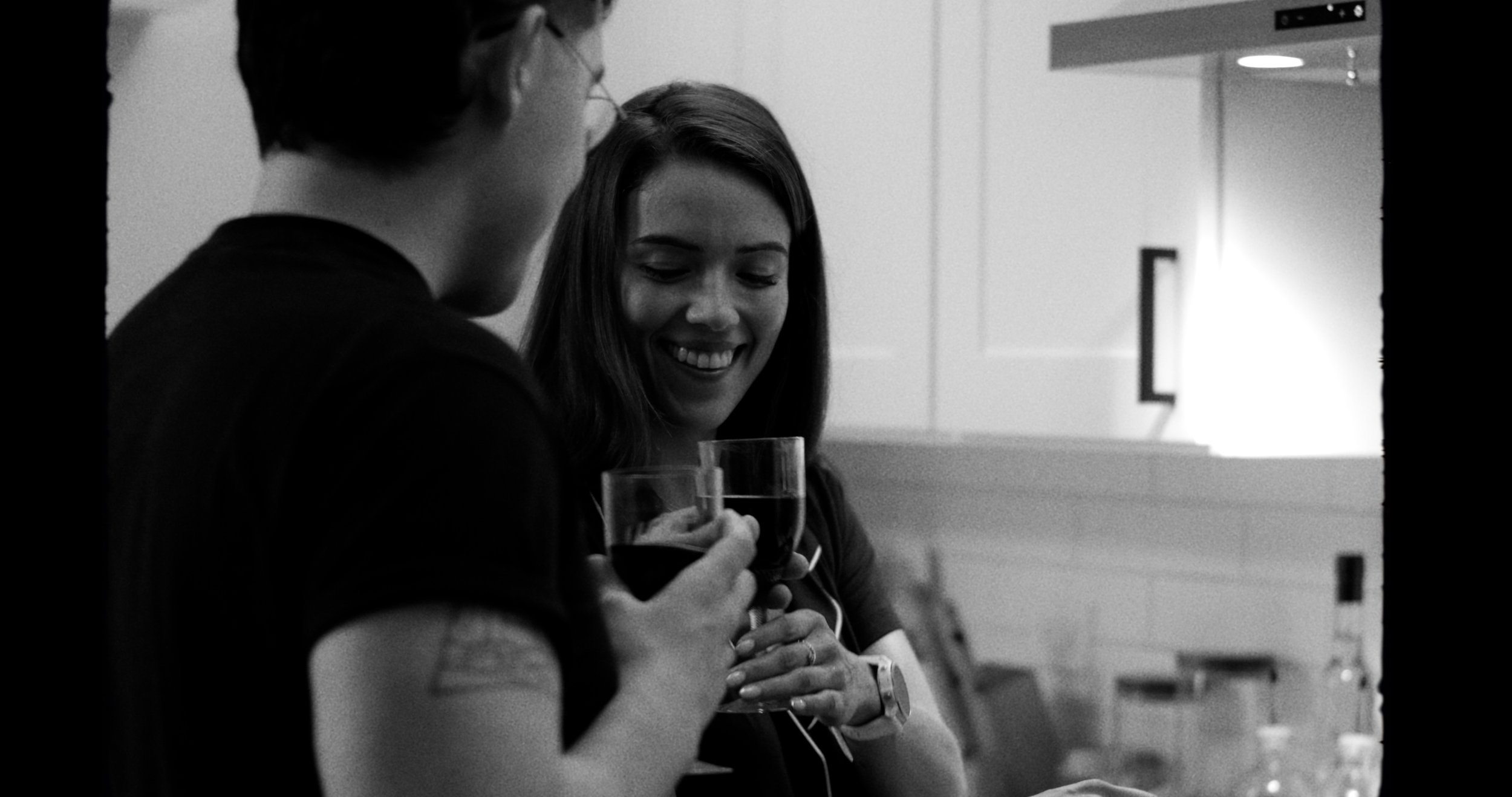 A black and white photo of two people smiling and holding glasses of wine in a kitchen.
