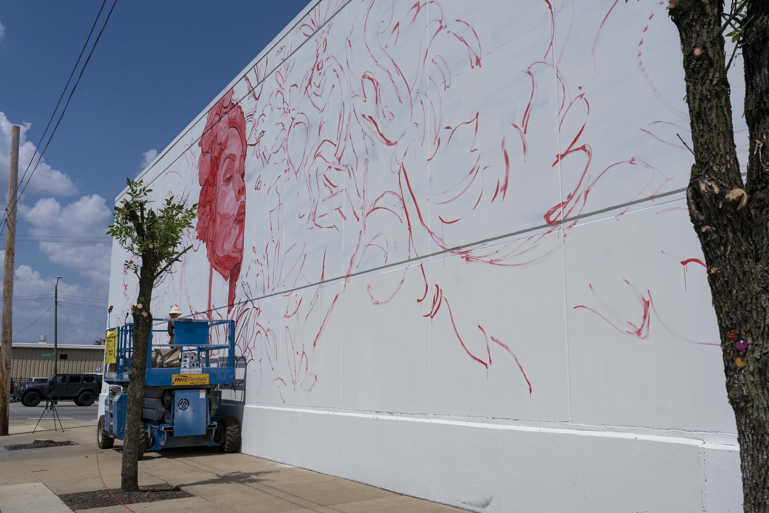 Artist working on a large mural in progress on the side of a building. The mural features red outlines and a partially completed portrait. A person on a blue lift is actively painting. Trees and a sidewalk are visible in the foreground under a blue s