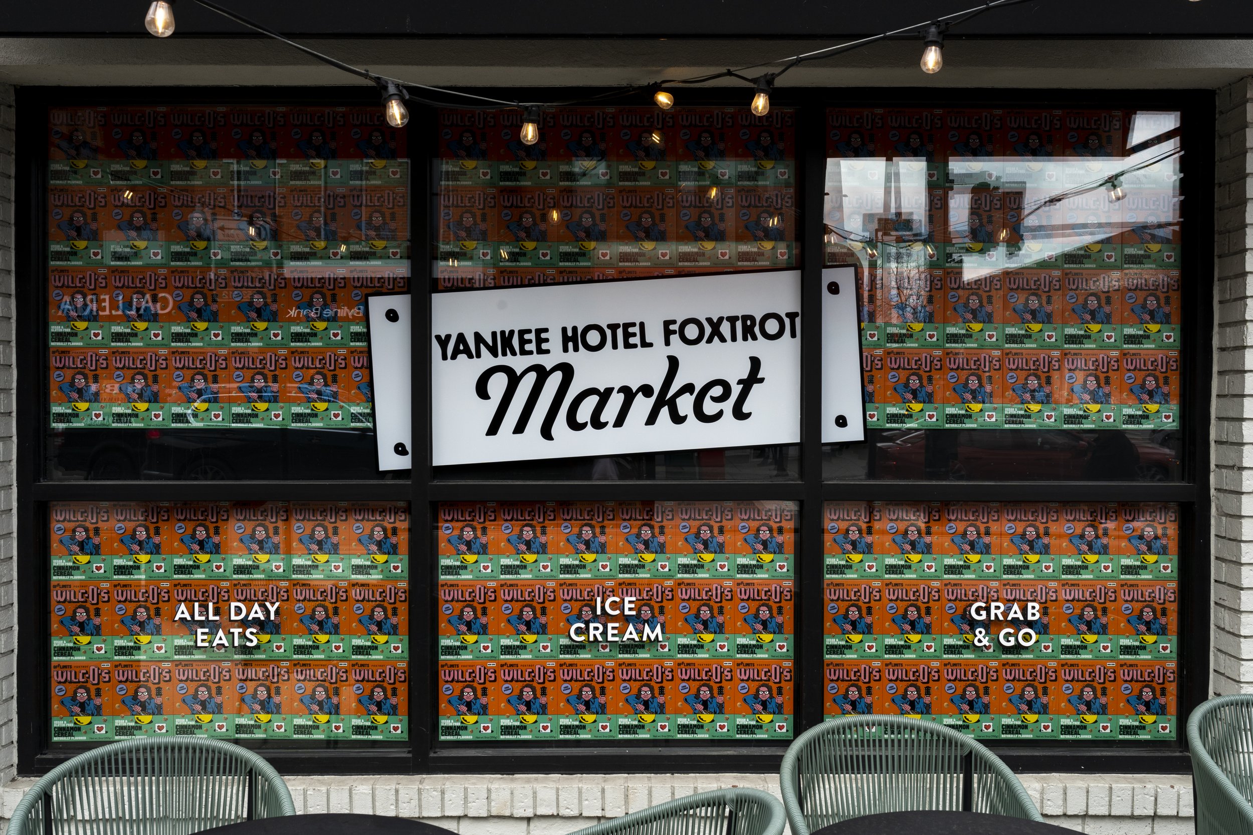 Storefront of Yankee Hotel Foxtrot Market with a white sign displaying the name. Windows decorated with colorful posters of Uncle J’s. Text on windows reads: All Day Eats, Ice Cream, Grab & Go. Outdoor seating includes green chairs and a table. Overh