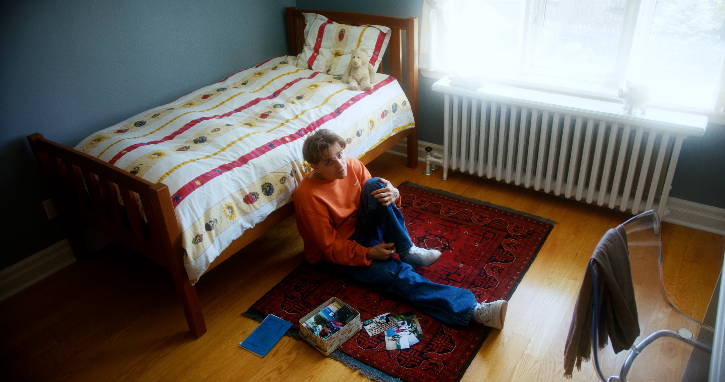 Person sitting on a red rug in a bedroom next to a bed with a colorful quilt, a small box with photos, and a notebook
