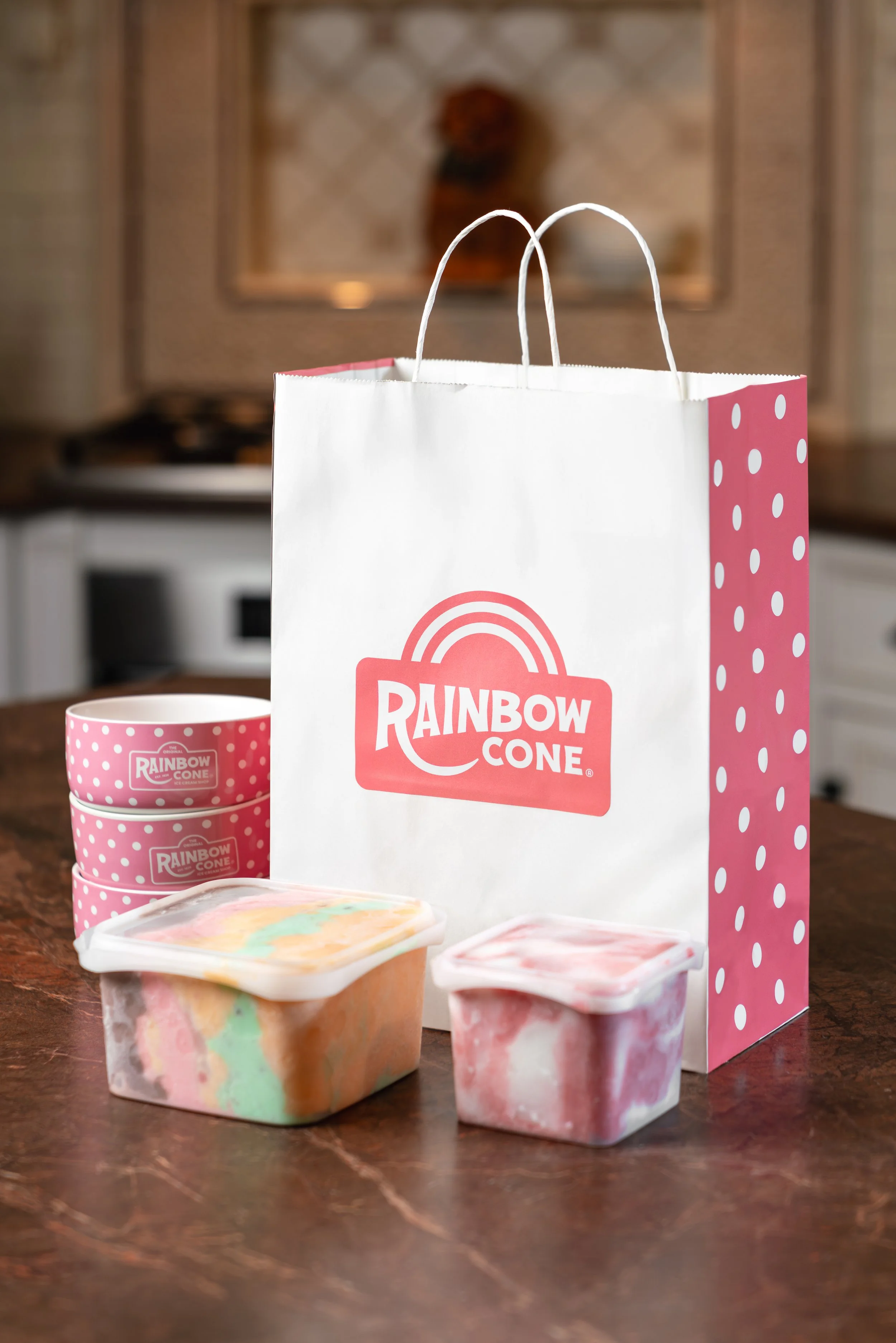 A Rainbow Cone shopping bag on a kitchen countertop with two tubs of rainbow-colored ice cream and three pink Rainbow Cone branded bowls stacked next to it.