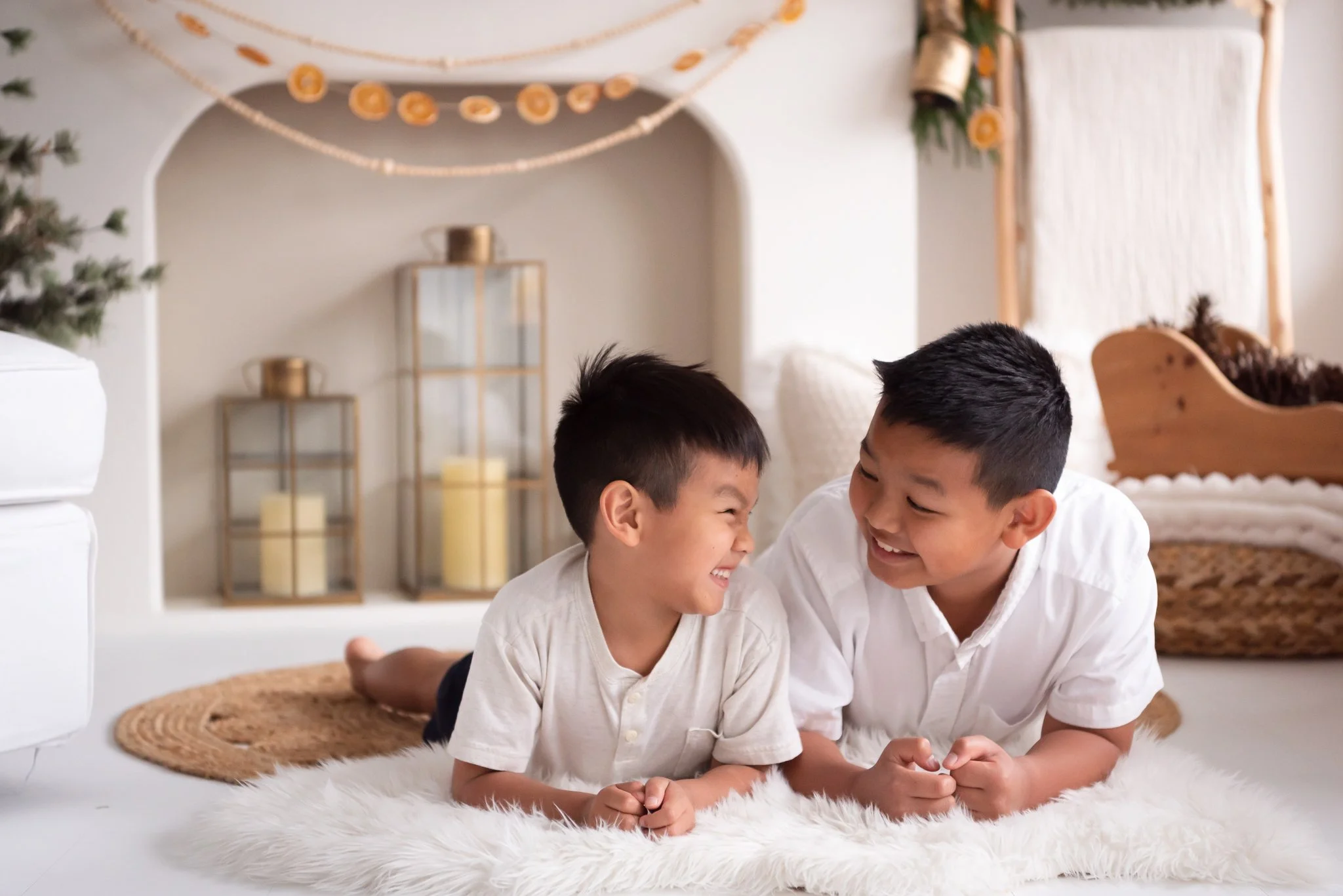 two brothers laying on the floor in front of holiday fireplace set. boys in white shirts and looking at each other grinning. background has candles, evergreen tree, strung oranges, and white fireplace.