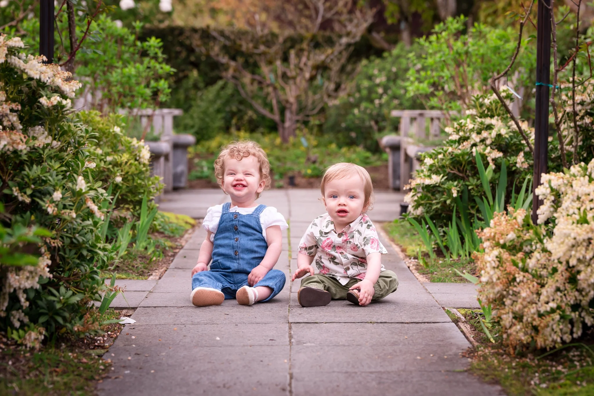 1 year old twin girl and boy sitting next to each other on a concrete path with green and flowering bushes around them