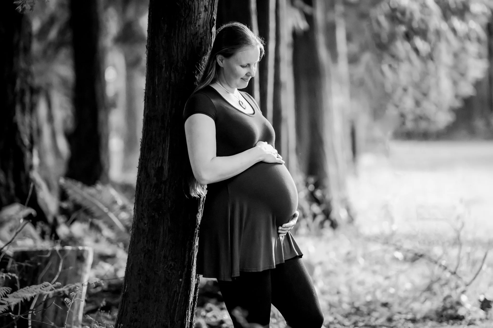 black and white image of Lindsay Eickhoff, photographer at Eickhoff Photography, leaning against an evergreen tree looking down at her pregnant belly in an Edmonds park for maternity photos