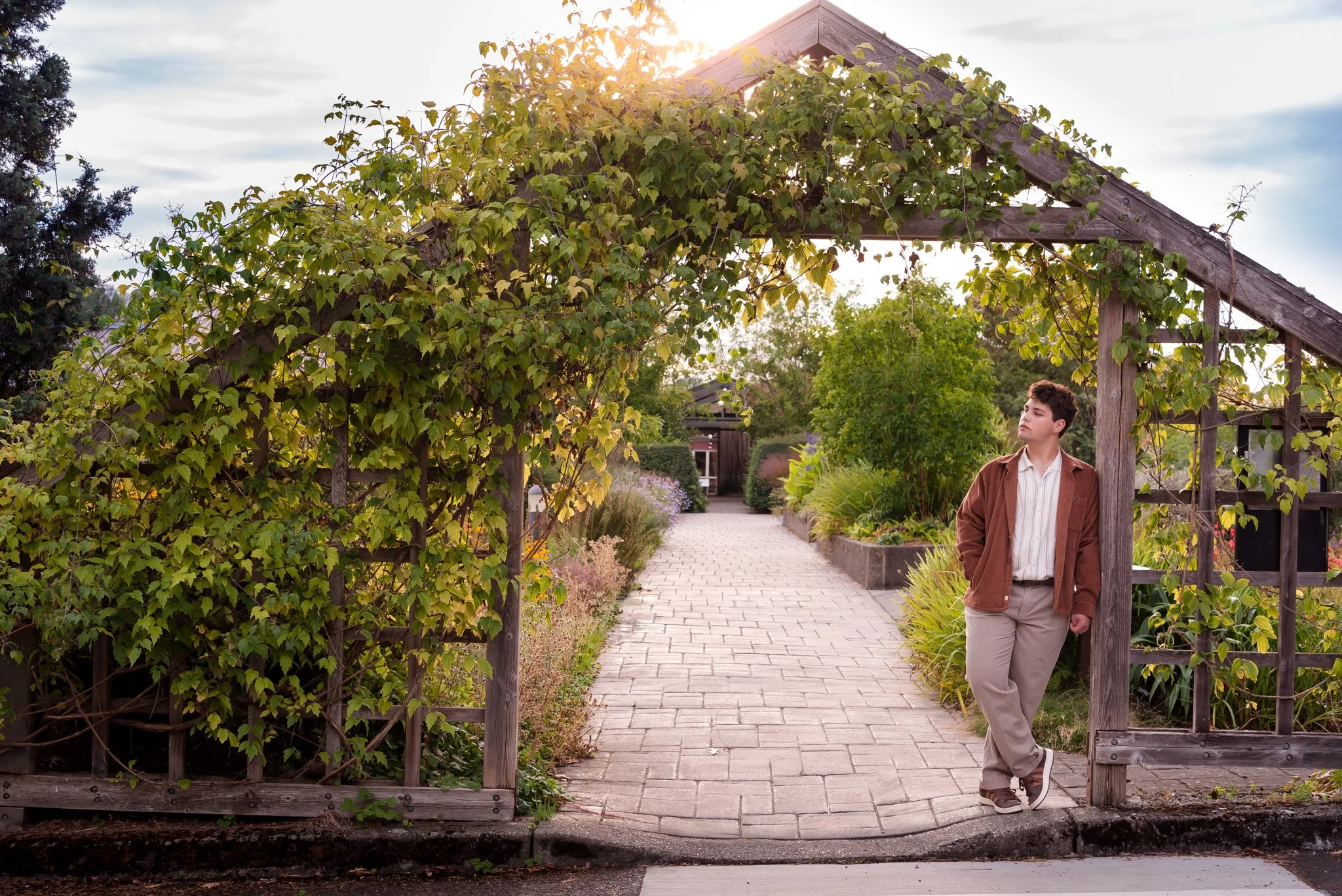 Senior boy leaning against trellised entrance gate covered in vines with garden behind him. He is looking off to the side with a sunflare above the vines and a cloudy sky.