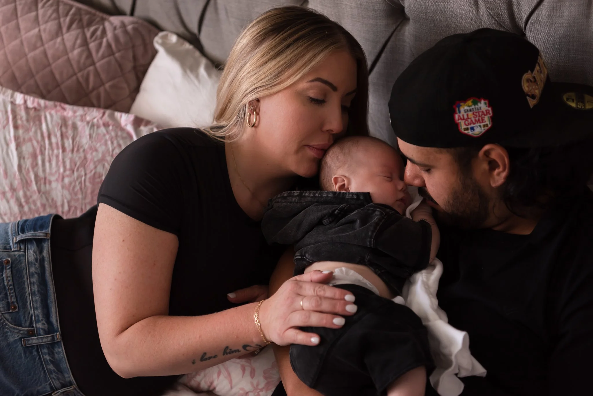 newborn baby boy laying in bed with mom and dad, all dressed in black and jeans with mom kissing baby's head and dad nuzzling his nose.