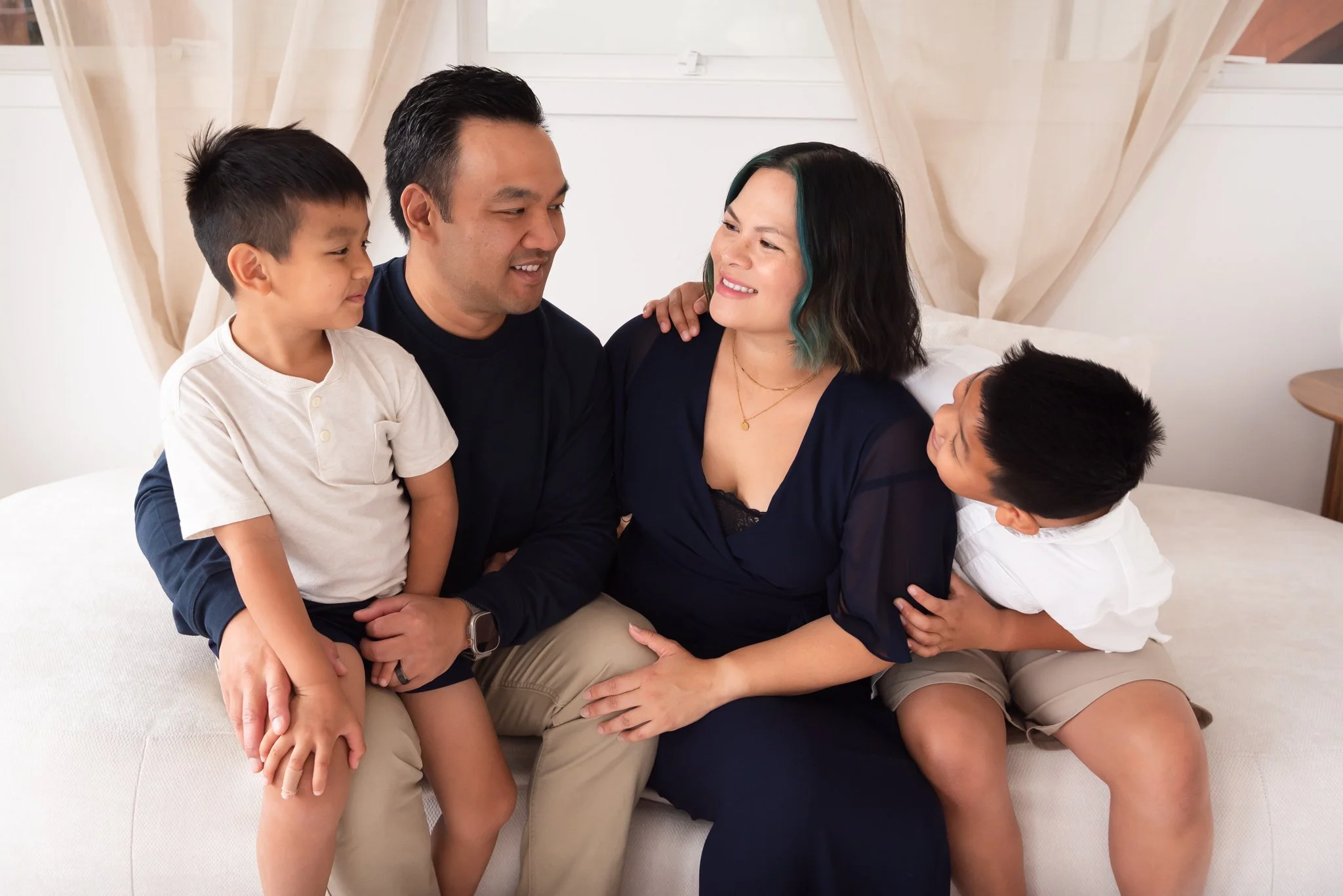 Family of four sitting on a cream colored couch in a studio living room with navy, khaki, and white clothing. Family smiling at each other.