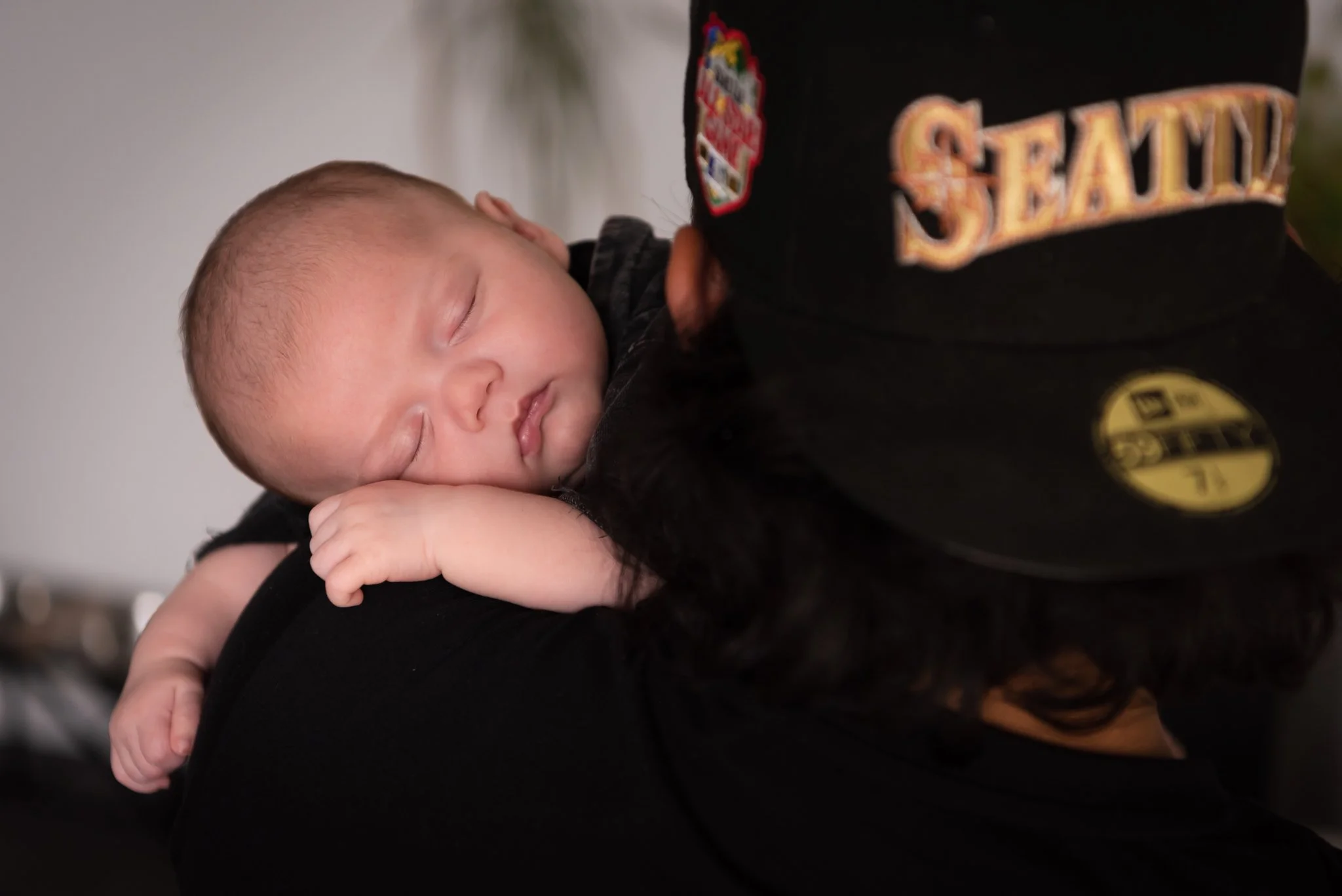 looking at the back of dad in black shirt and black and gold Seattle Mariners hat holding baby boy up on his shoulder. Baby is sleeping while snuggled on dad.